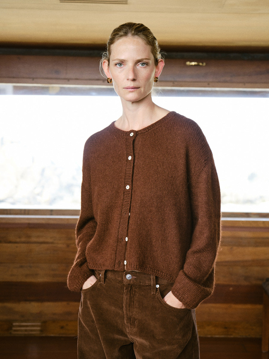 A woman wearing a brown button-front knit cardigan and matching brown corduroy pants, standing indoors in front of a bright window with wood paneling.