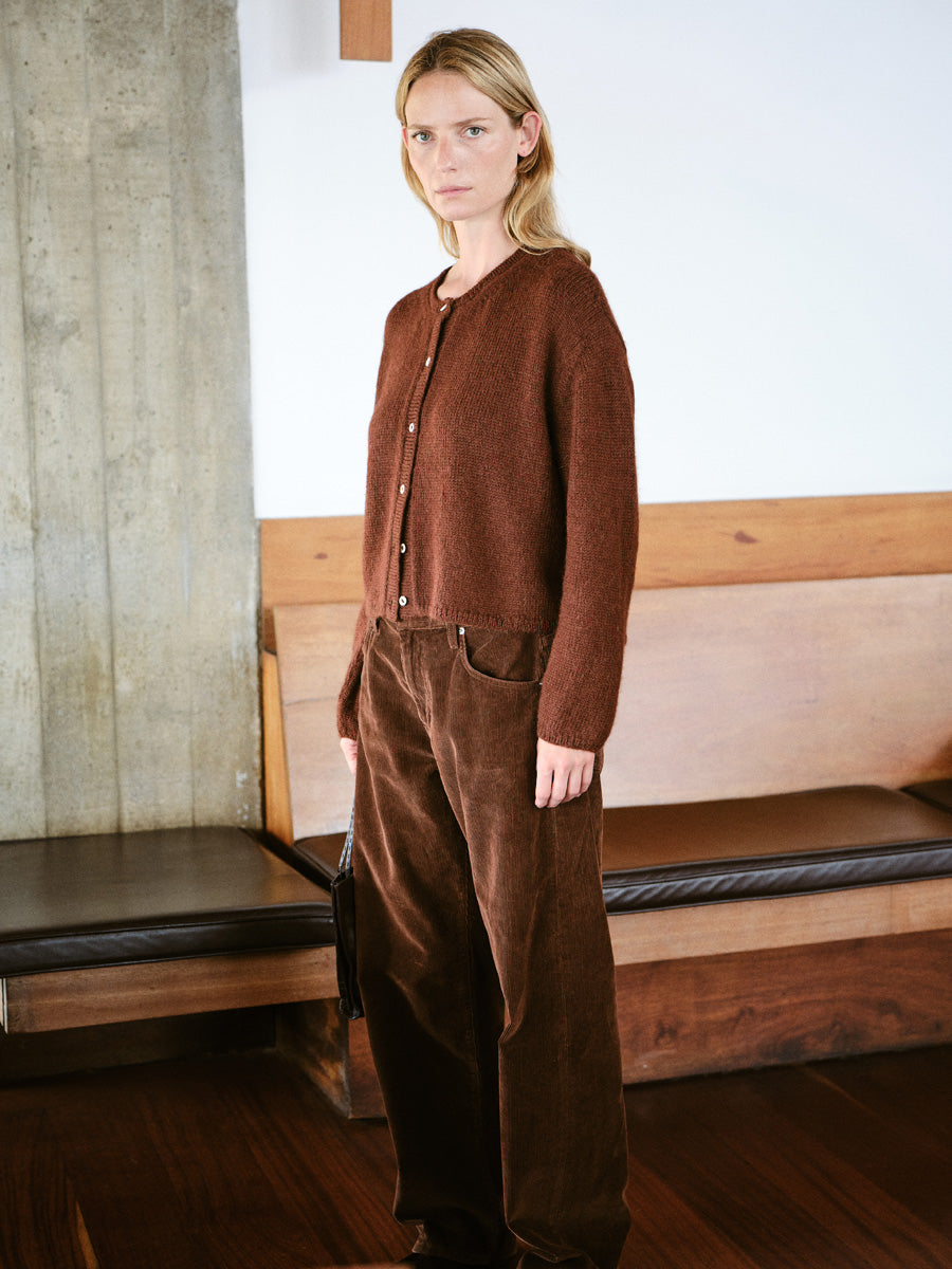 A woman stands indoors wearing a brown knit button-front cardigan with matching brown corduroy pants. The setting features a concrete pillar, wooden bench, and dark wood floor.