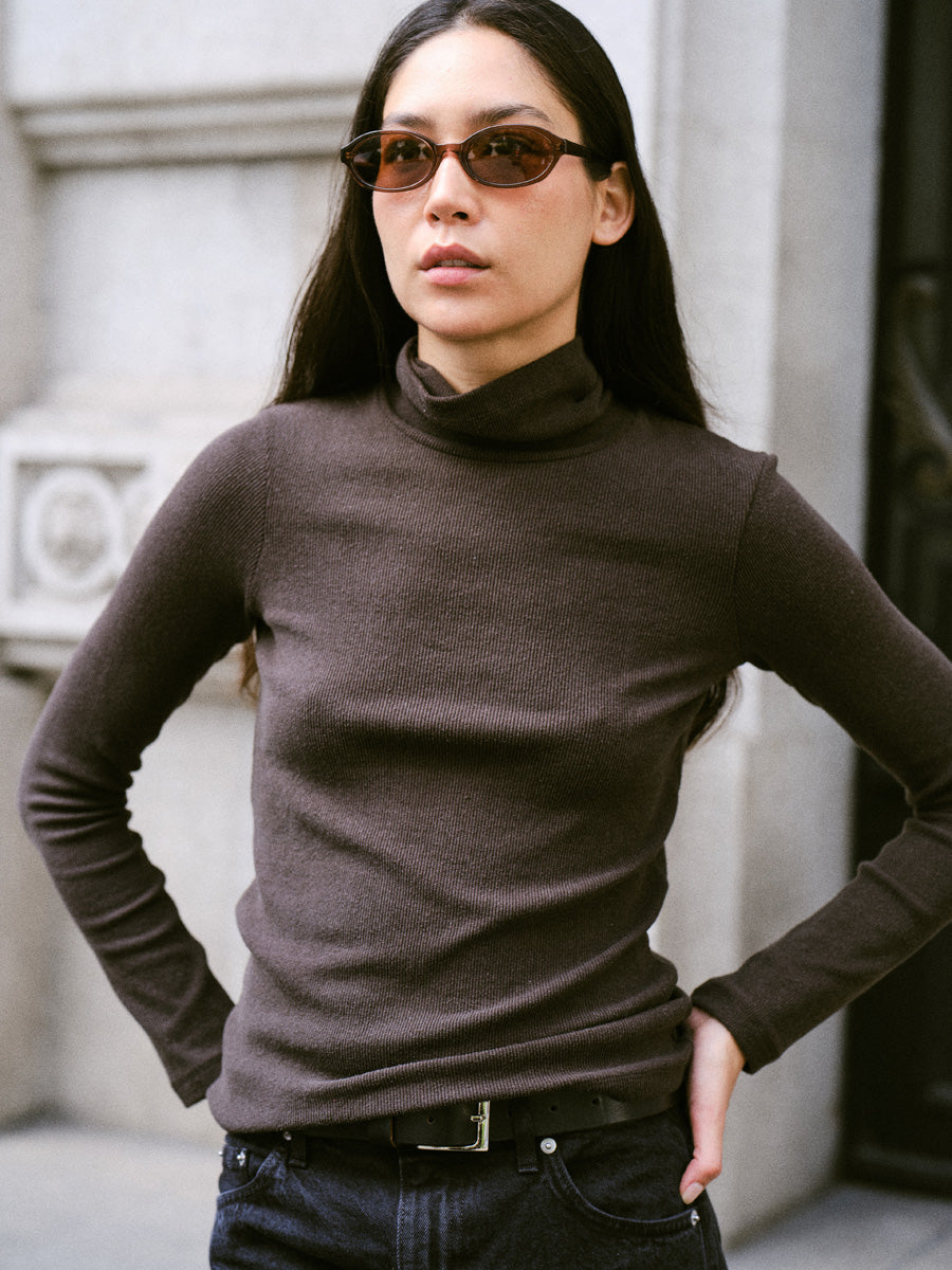 A woman standing on the street with sunglasses and wearing a long-sleeved dark brown turtleneck and black jeans.
