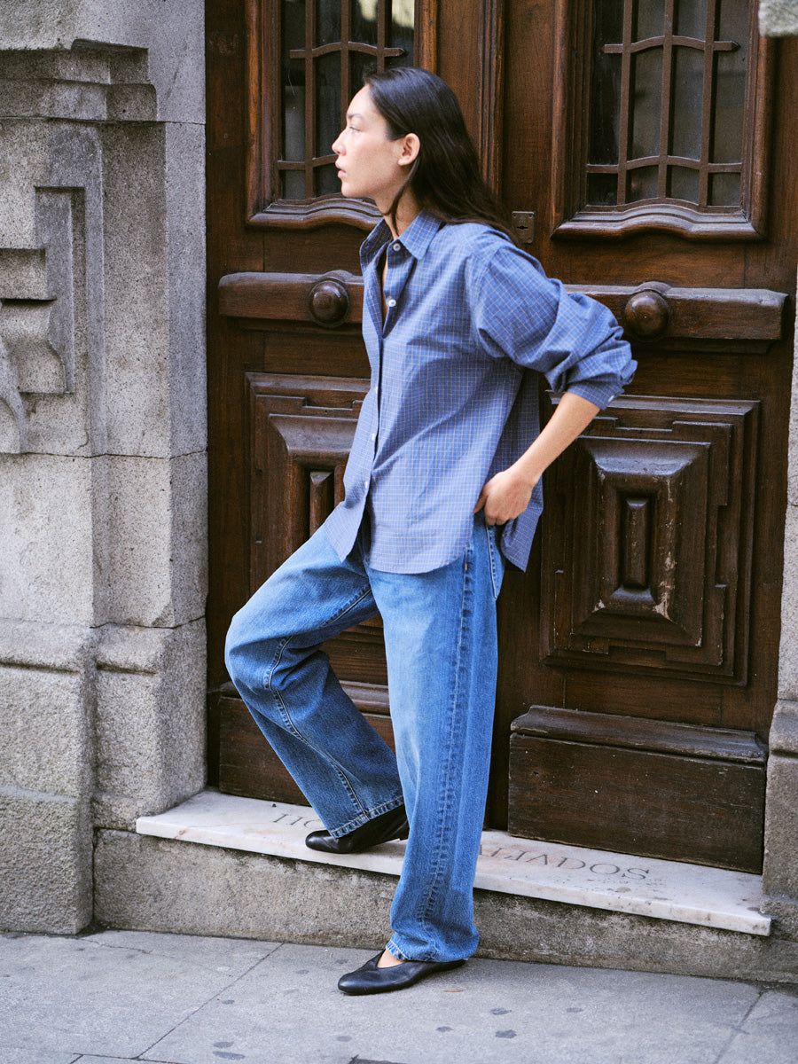 A woman wearing a light blue button-down shirt and wide-leg blue jeans stands on stone steps in front of a dark wooden door.