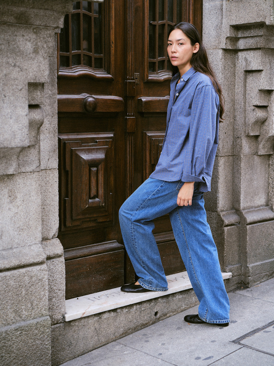 A woman wearing a blue button-up shirt and wide-leg blue jeans stands on a stone doorstep next to a dark wooden door.