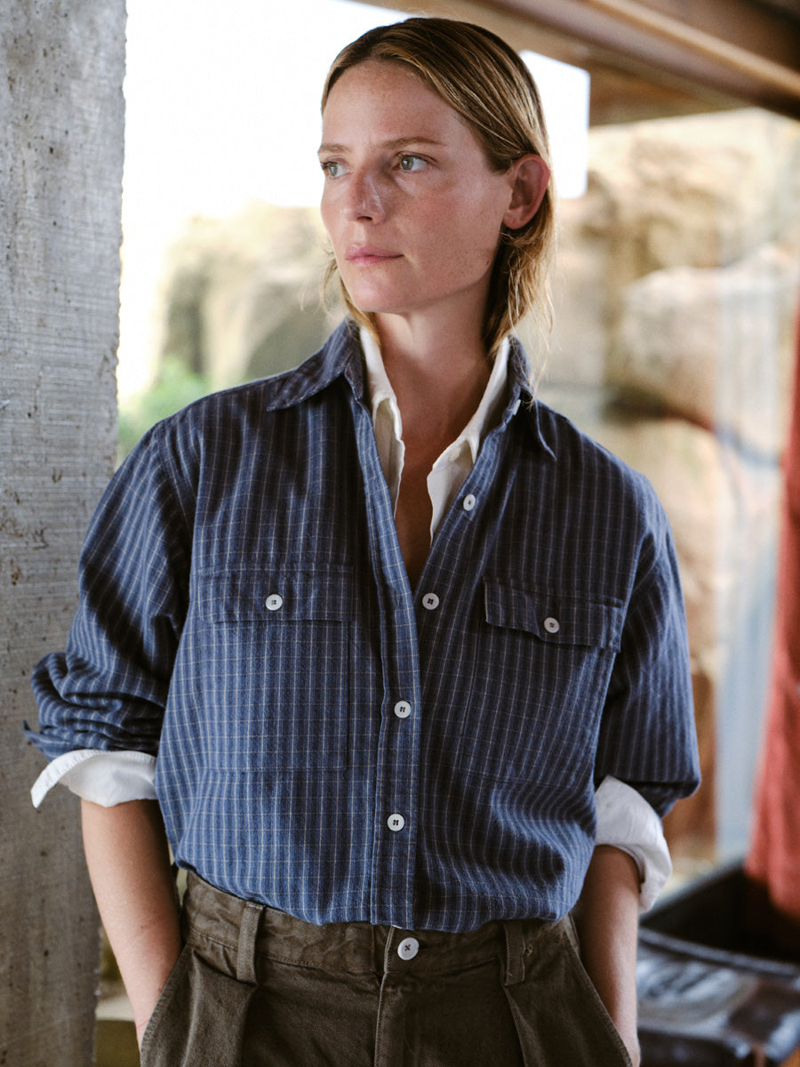 A woman wearing a blue and white vertically striped button-up shirt over a white undershirt, paired with brown trousers, standing in a rustic stone-walled environment.