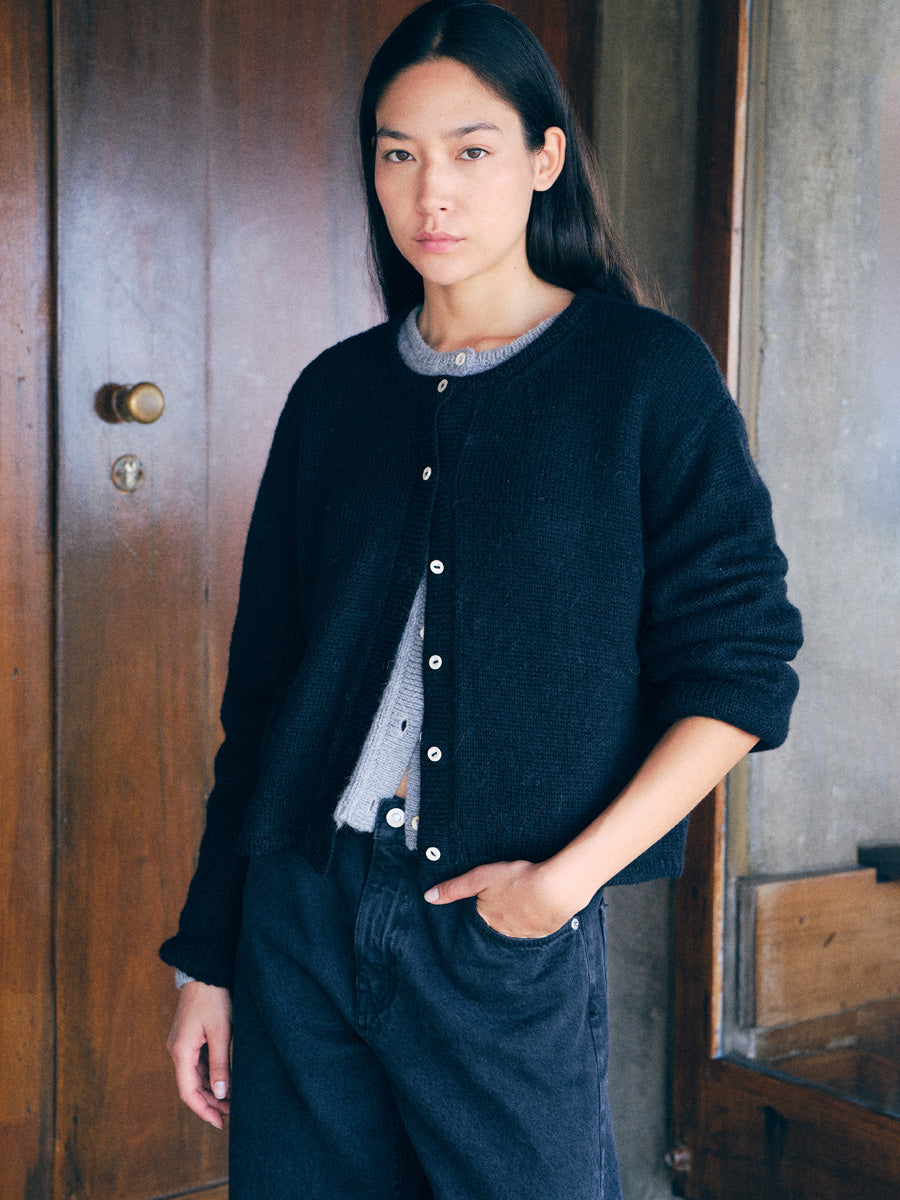 A woman wearing a black button-up cardigan over a gray knit top and dark jeans, standing in front of a rustic wooden door.