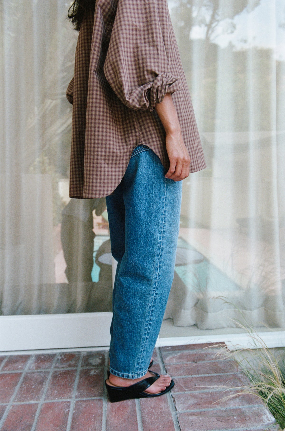 A person wearing an oversized brown and beige gingham shirt with ruffled sleeves and blue wide-leg jeans, standing on a brick patio in front of a window with sheer curtains.