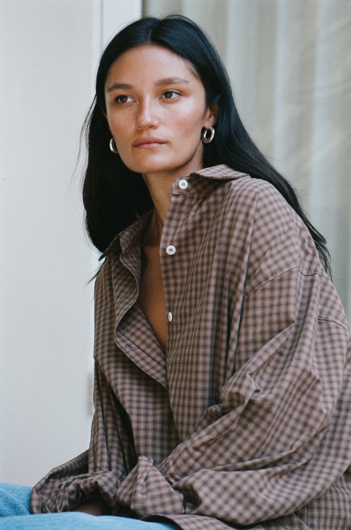 A woman with long dark hair wearing an oversized brown and beige gingham button-up shirt, sitting against a neutral light background.