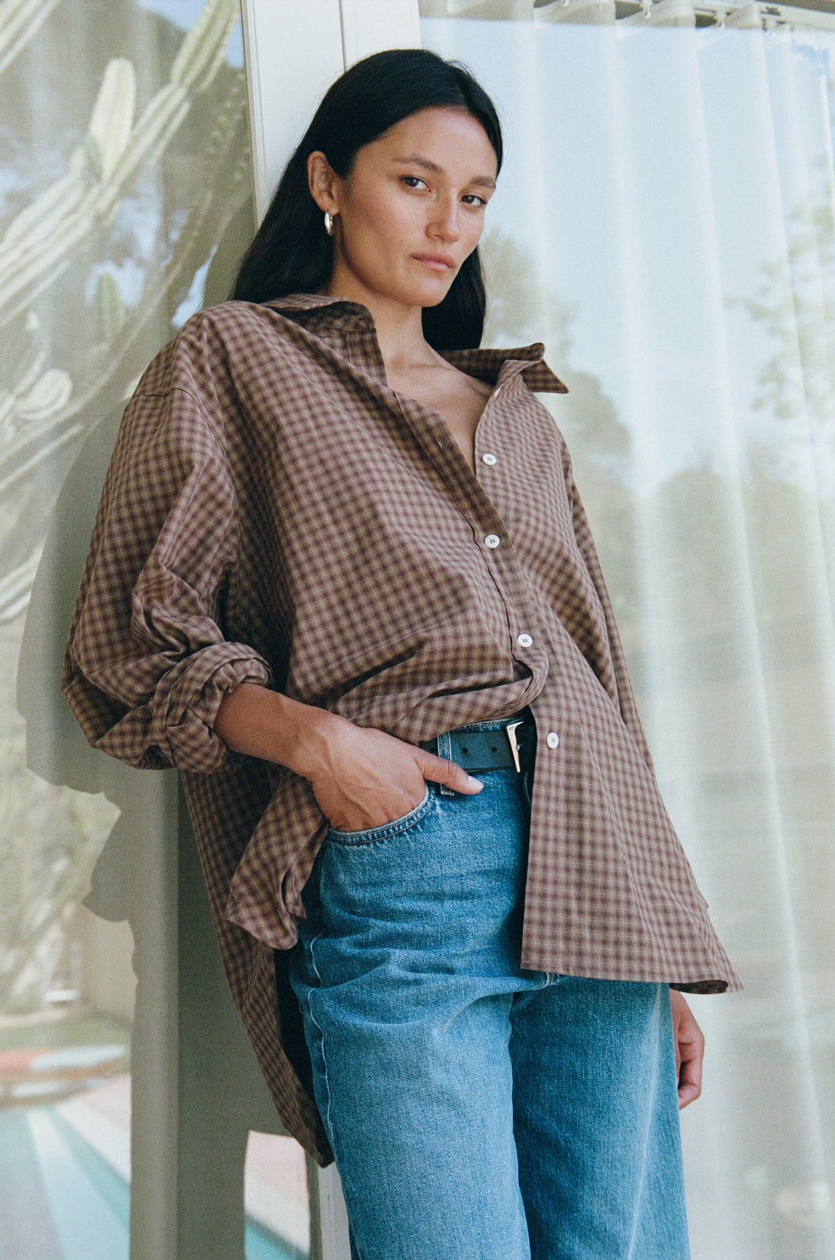 A woman wearing an oversized brown and beige gingham button-up shirt tucked into blue jeans, leaning against a window with sheer curtains.