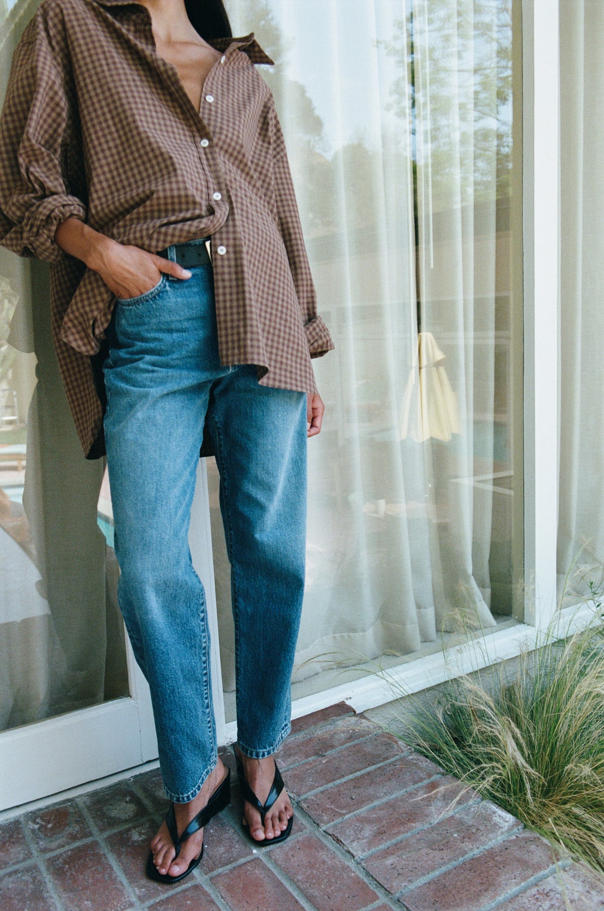 A person wearing a brown and beige gingham button-up shirt, blue straight-leg jeans, and black thong sandals, standing on a brick patio in front of a glass door with sheer curtains.