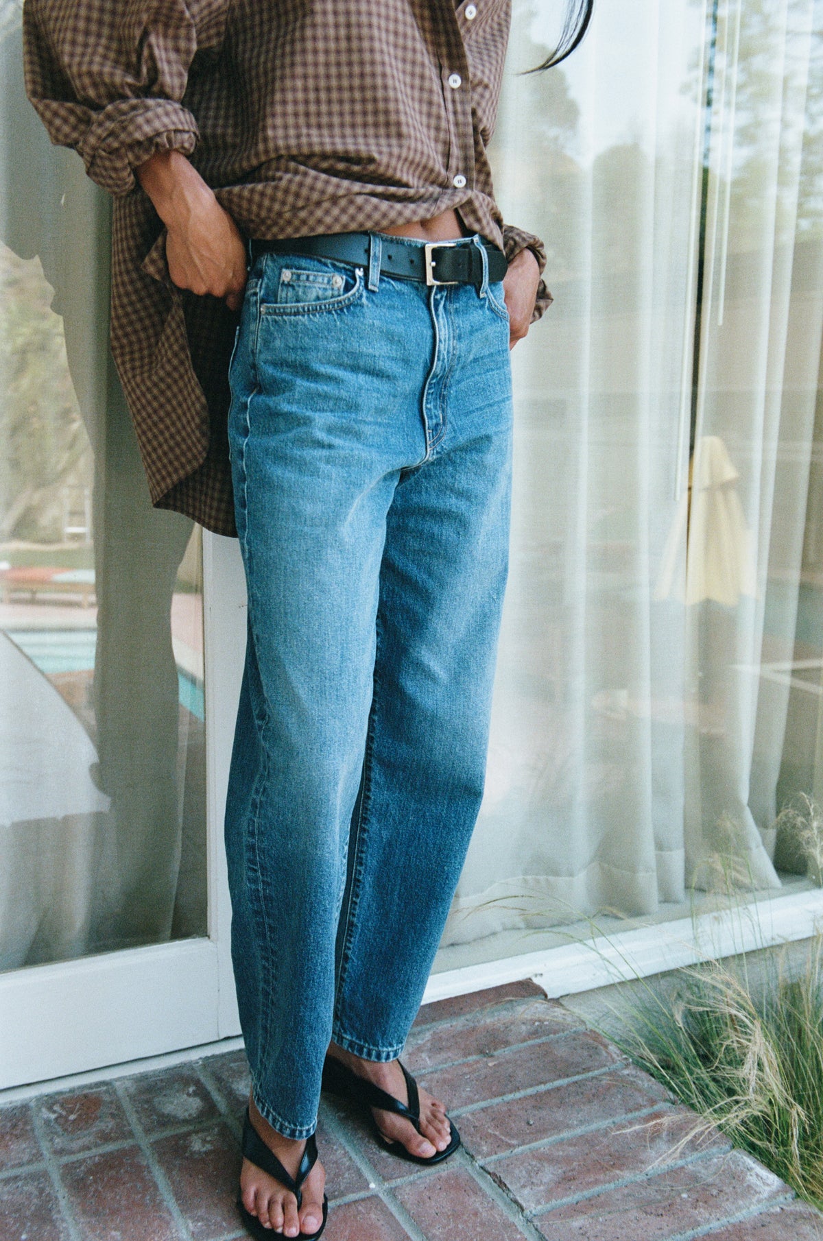 A person wearing medium-wash blue straight-leg jeans with a brown and beige gingham button-up shirt and black flip-flops, standing on a brick patio in front of a glass door with sheer curtains.