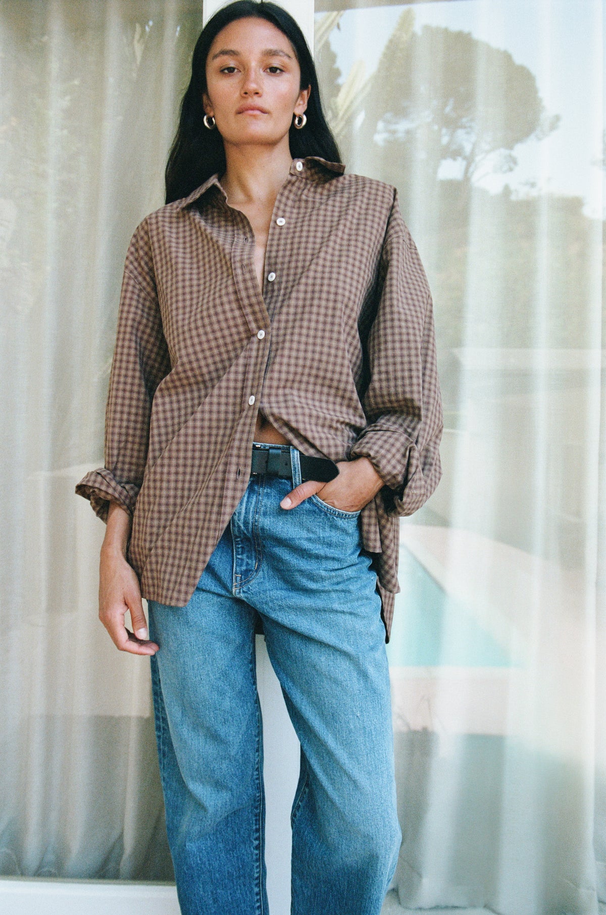 A woman wearing a brown and beige gingham button-up shirt with rolled sleeves and blue jeans, standing in front of sheer curtains.