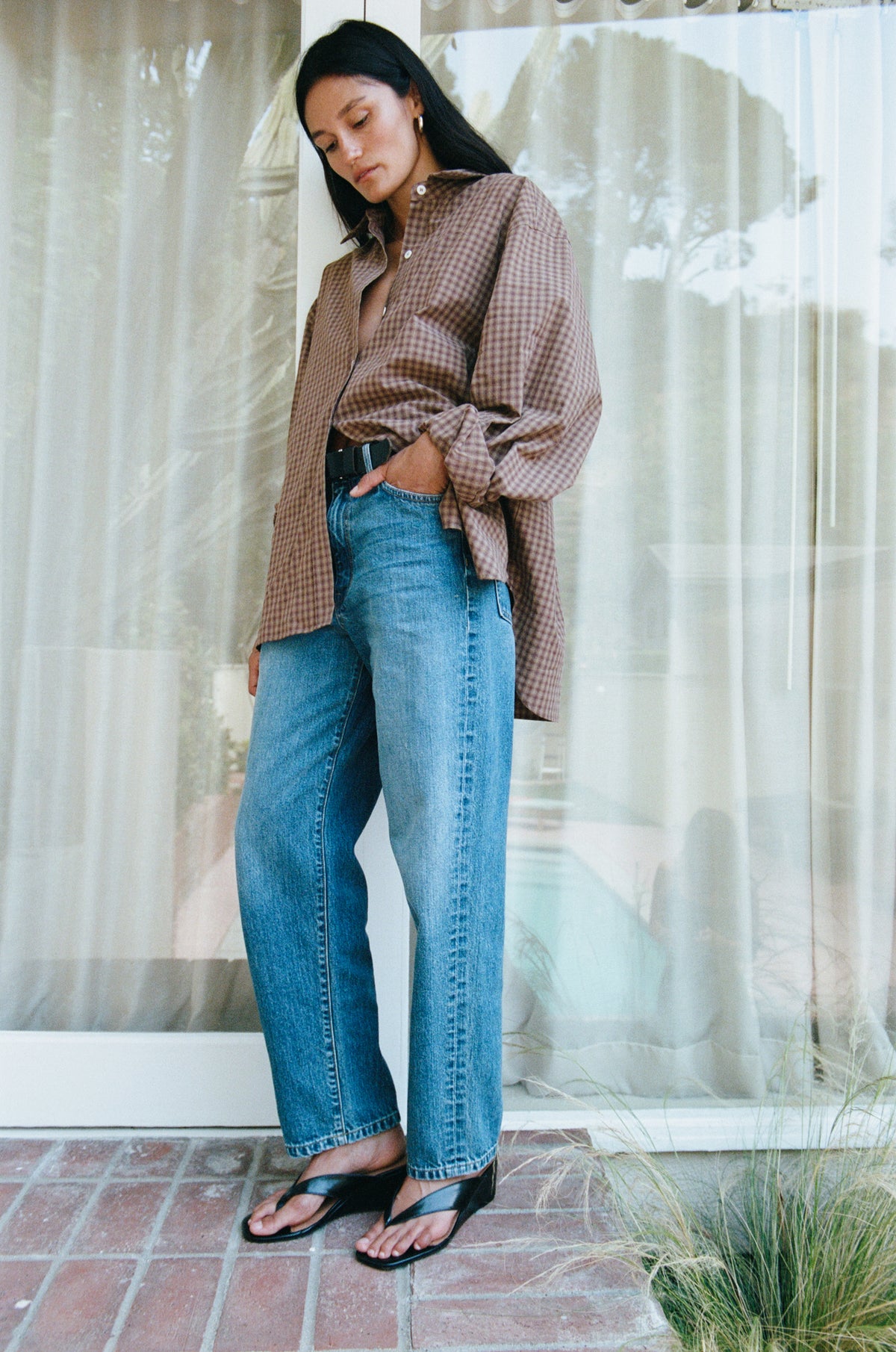 A woman stands outdoors in front of sheer curtains and glass doors, wearing an oversized brown-and-white gingham button-up shirt, blue wide-leg jeans, and black slide sandals.