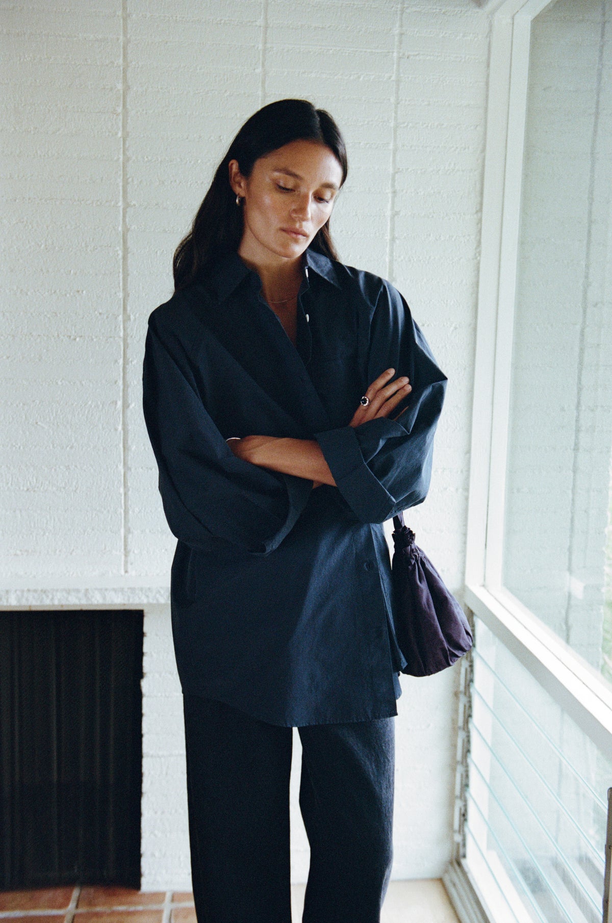 A woman stands indoors near a window wearing an oversized dark navy button-up shirt and matching dark pants, with a dark shoulder bag. The background shows a white brick wall and a window.