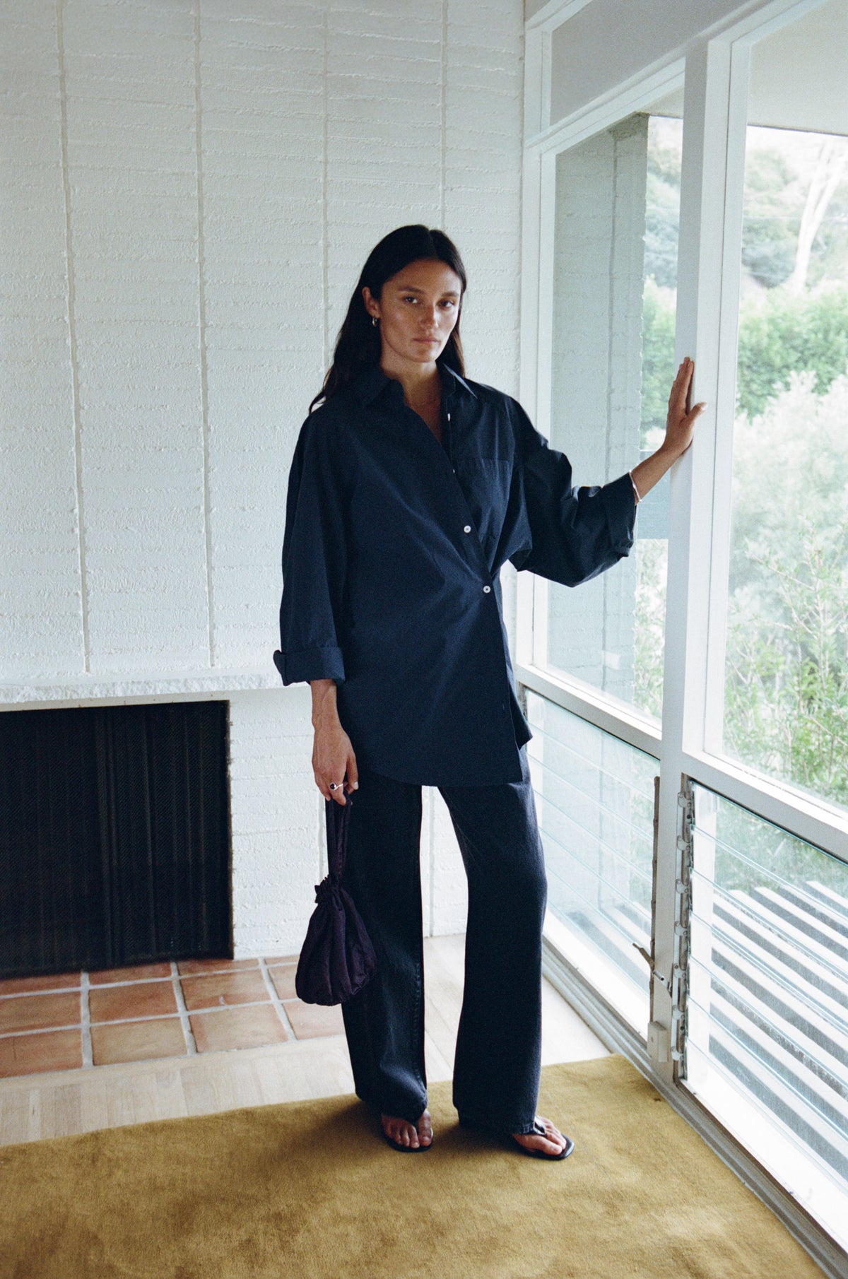 A woman stands indoors by large windows wearing a dark navy long-sleeve button-up shirt and matching wide-leg pants, holding a dark handbag. The room has white textured walls, a fireplace, and a tan rug.