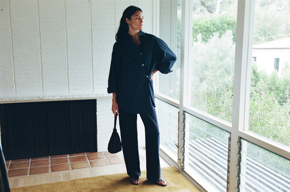 A woman stands by a large window in a minimalist room, wearing a dark navy blue oversized button-up shirt and matching wide-leg trousers, holding a black handbag.