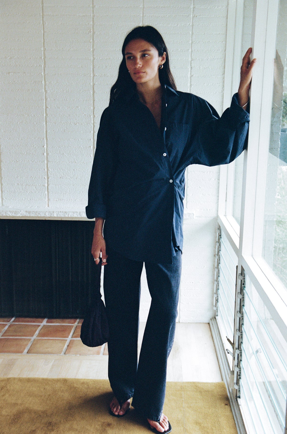 A woman stands by a window wearing a dark navy blue button-up shirt and matching wide-leg pants, holding a small dark handbag.
