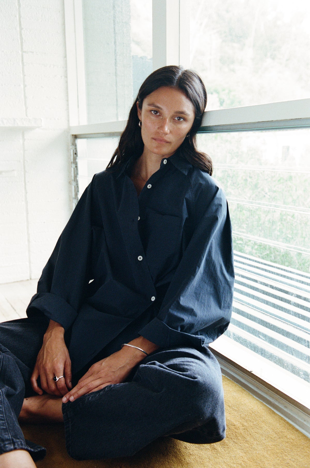 A woman sits on a wooden floor in front of a large window, wearing a dark navy blue long-sleeve button-up shirt and matching dark pants.