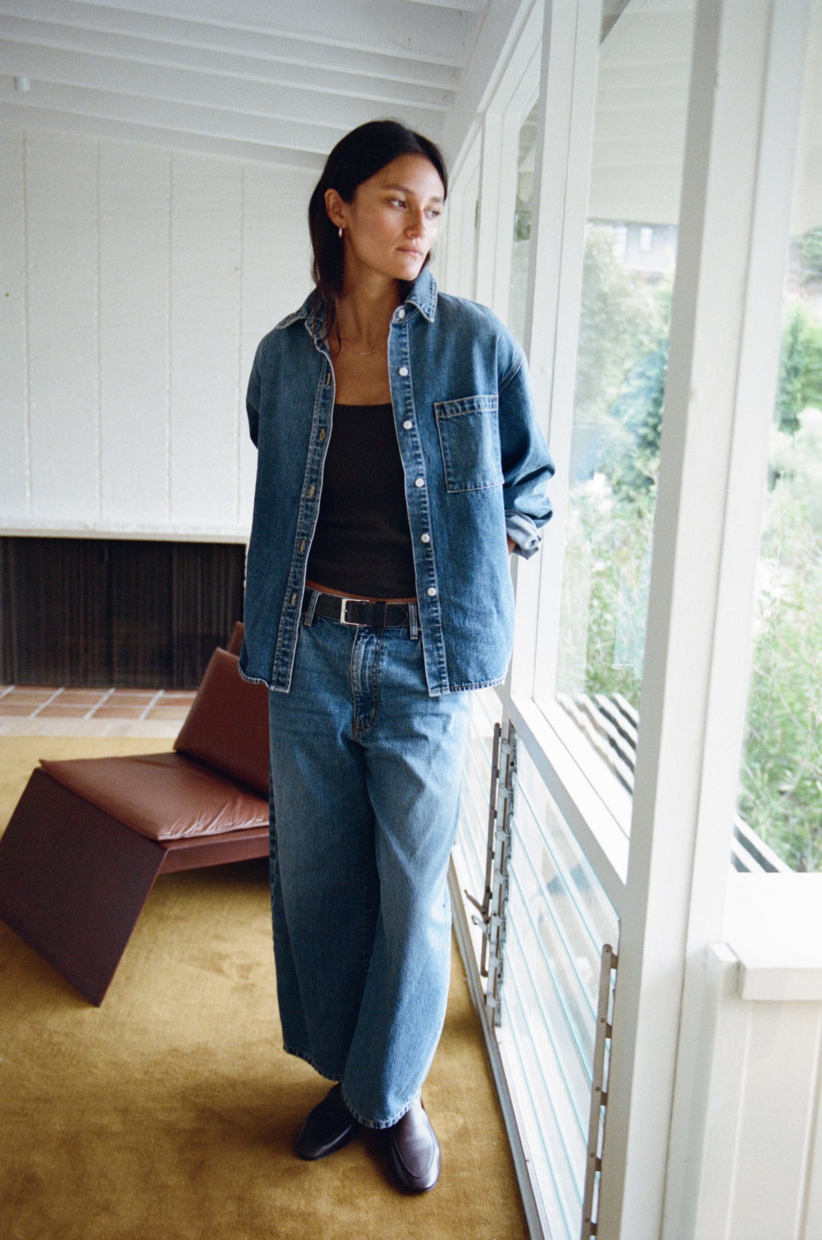 A woman stands on a porch wearing a blue denim shirt and matching wide-leg jeans over a black top, with a brown leather chair and large windows in the background.