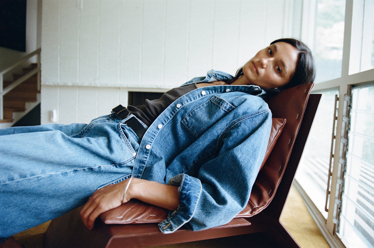 A woman reclining in a brown leather chair, wearing a blue denim shirt and matching wide-leg jeans in a modern interior setting.