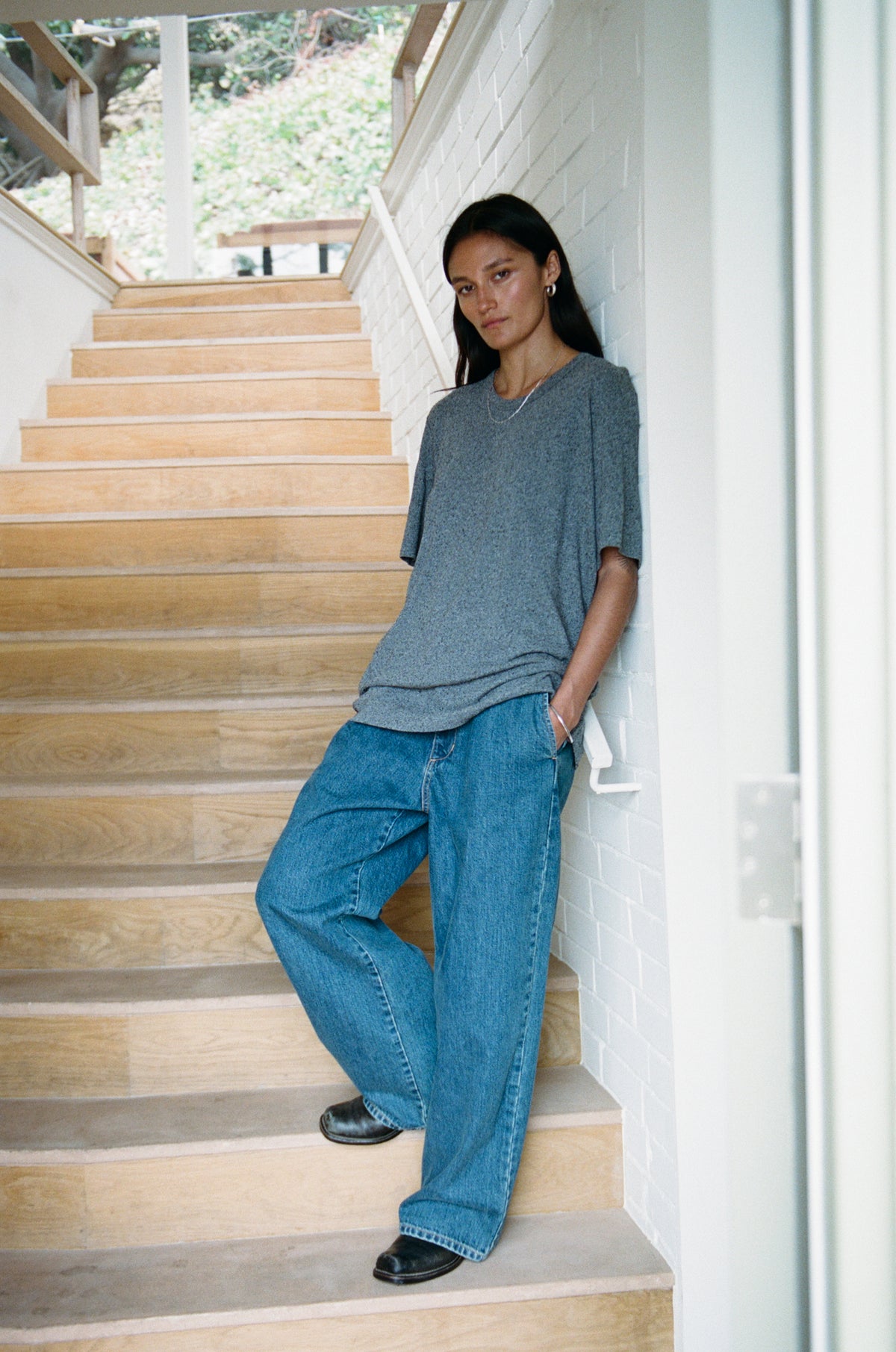 A woman wearing a loose gray t-shirt and wide-leg blue jeans leans against a white brick wall on a wooden staircase.