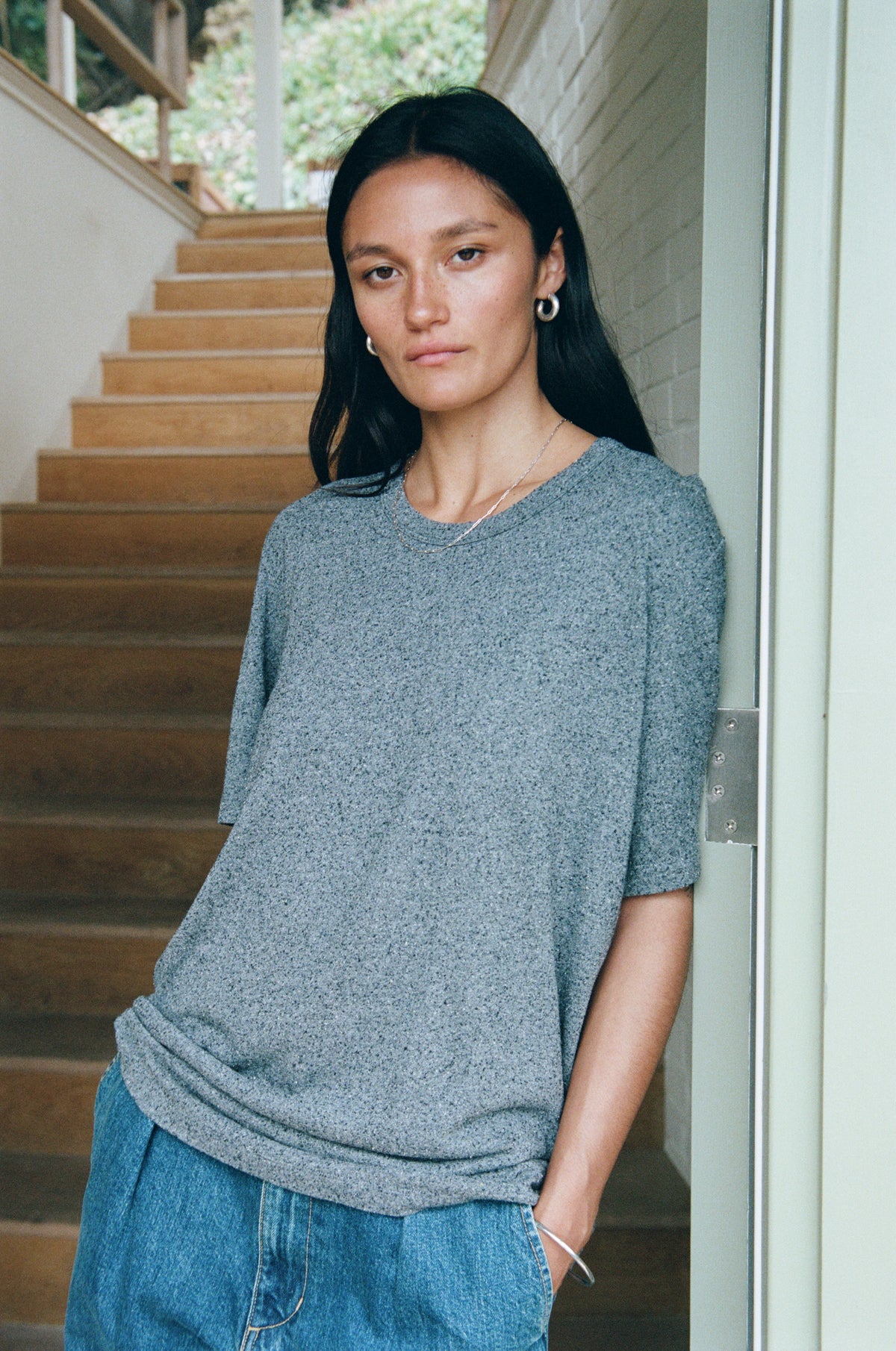 A woman with long black hair stands in a doorway wearing a loose-fitting, short-sleeve heathered gray knit top and blue jeans, with wooden stairs in the background.