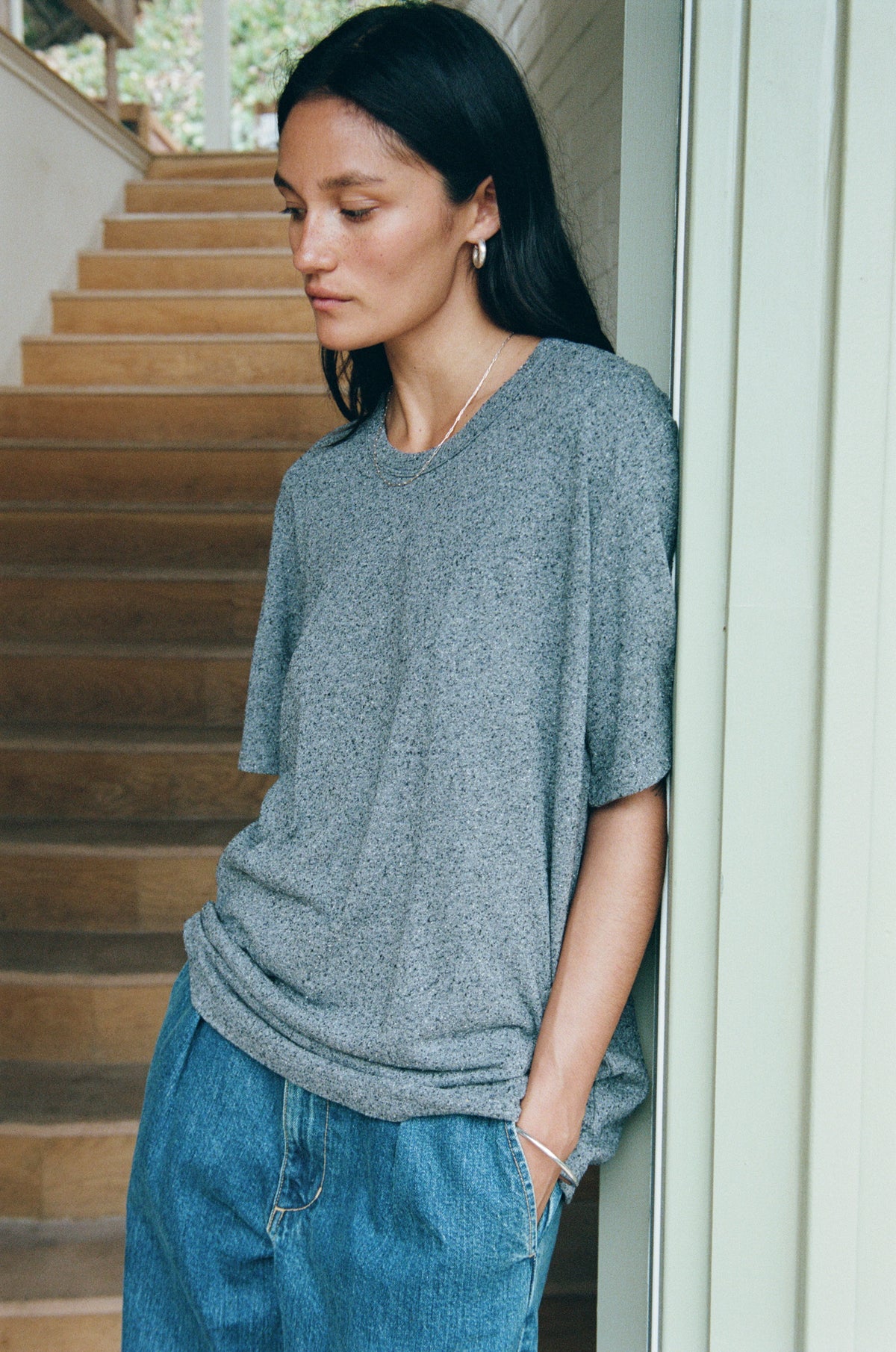 A woman wearing a heather gray short-sleeve knit top and blue jeans, leaning against a white doorframe next to wooden stairs.