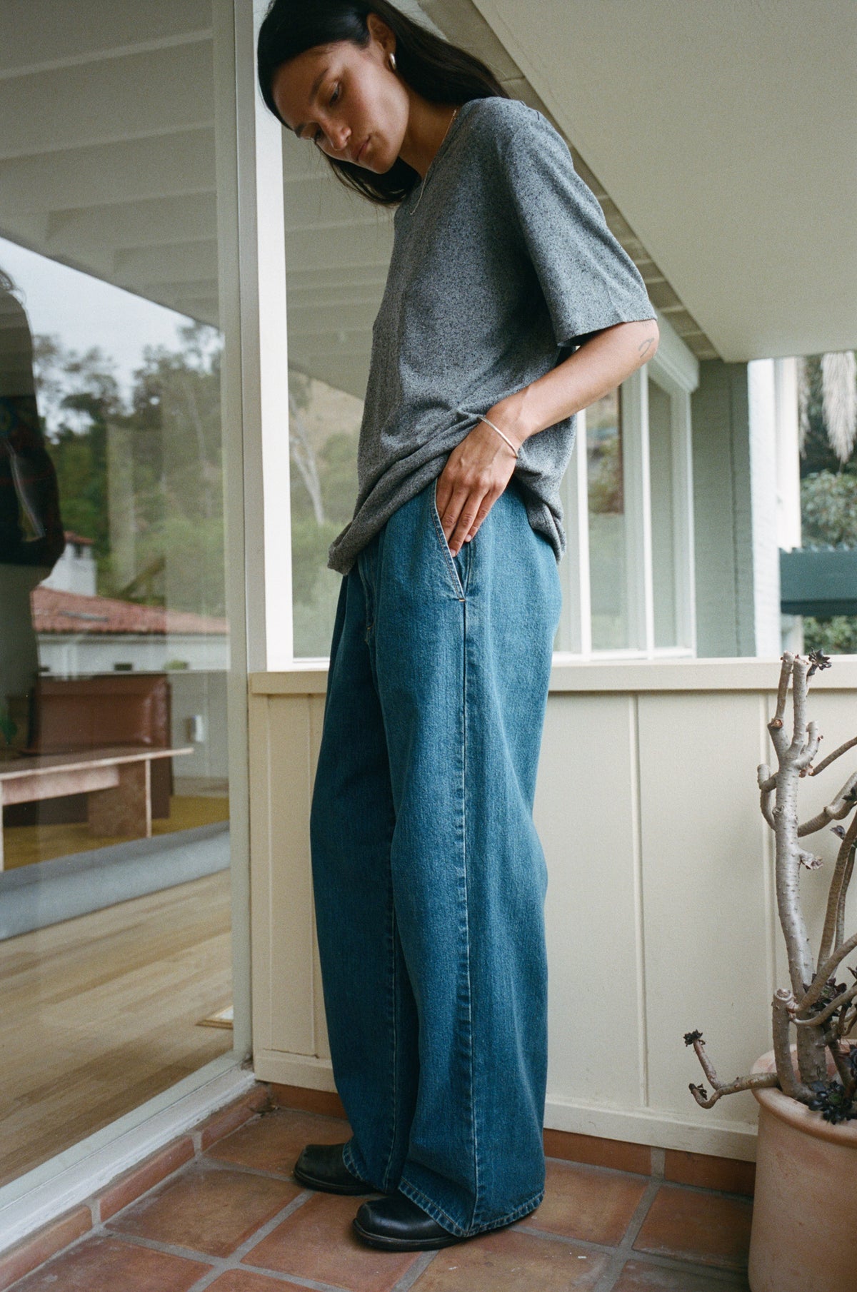 A woman wearing a gray oversized t-shirt and wide-leg blue denim jeans, standing on a tiled porch with a glass door behind her.