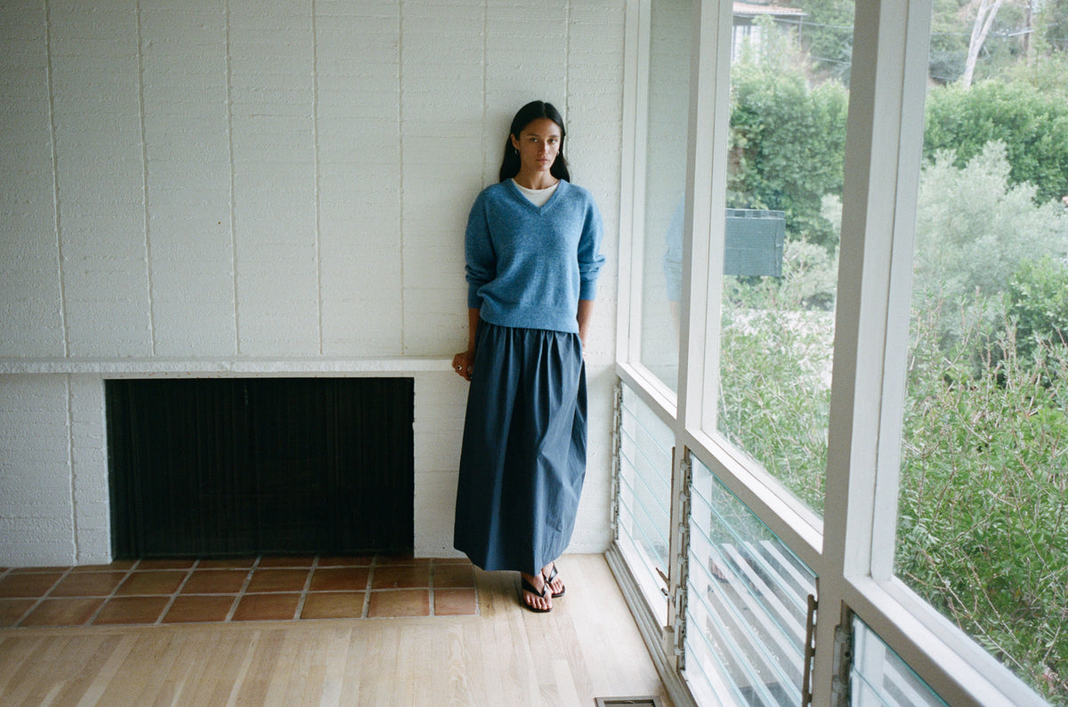 A woman stands on a sunlit porch wearing a blue long-sleeve knit sweater and a dark navy maxi skirt, with a white-paneled wall and large windows behind her.