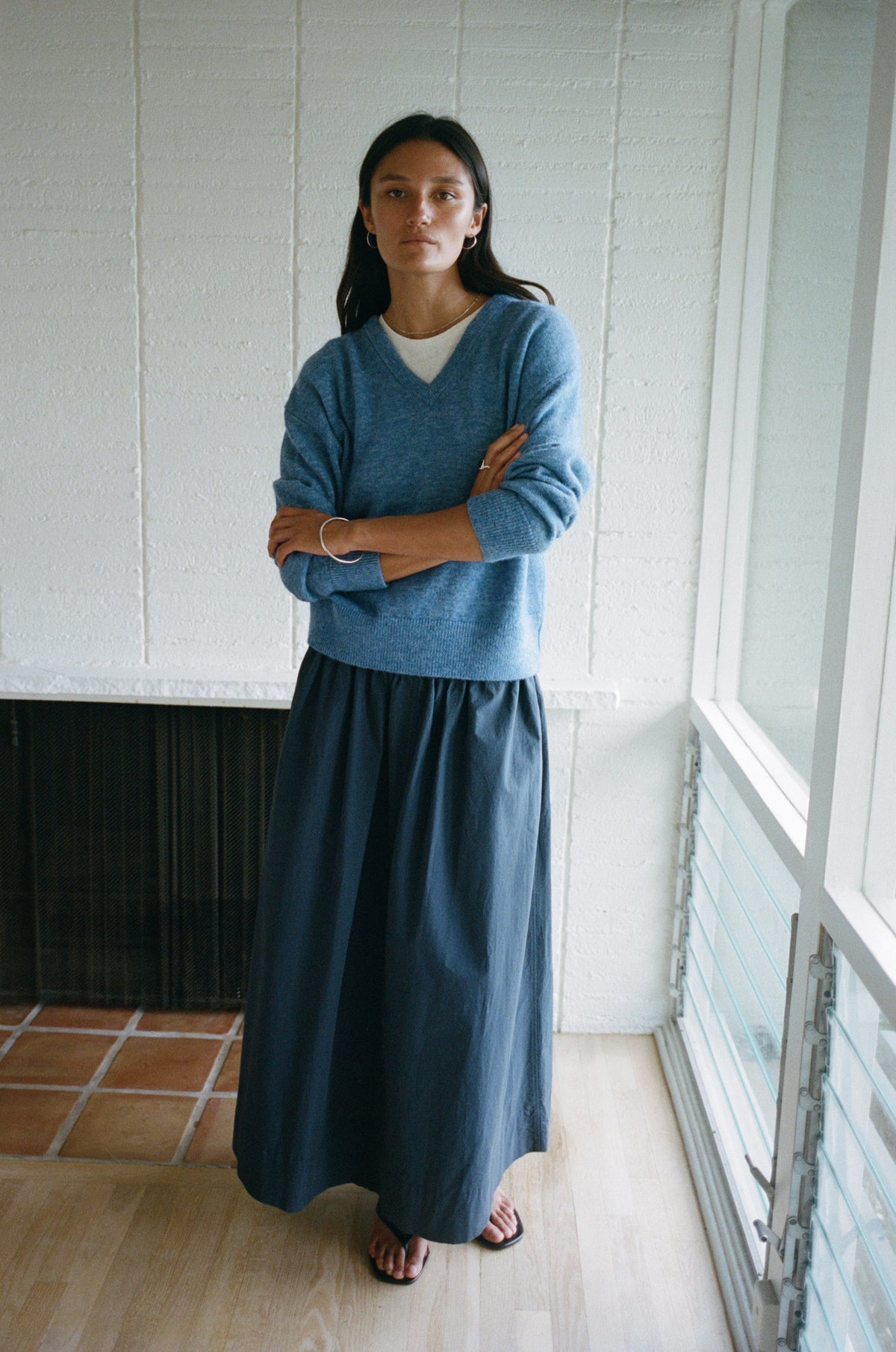 A woman stands indoors with her arms crossed, wearing a blue long-sleeve knit sweater over a white top and a dark blue maxi skirt. The setting has a white paneled wall, a dark fireplace opening, and a window with light coming in from the right.