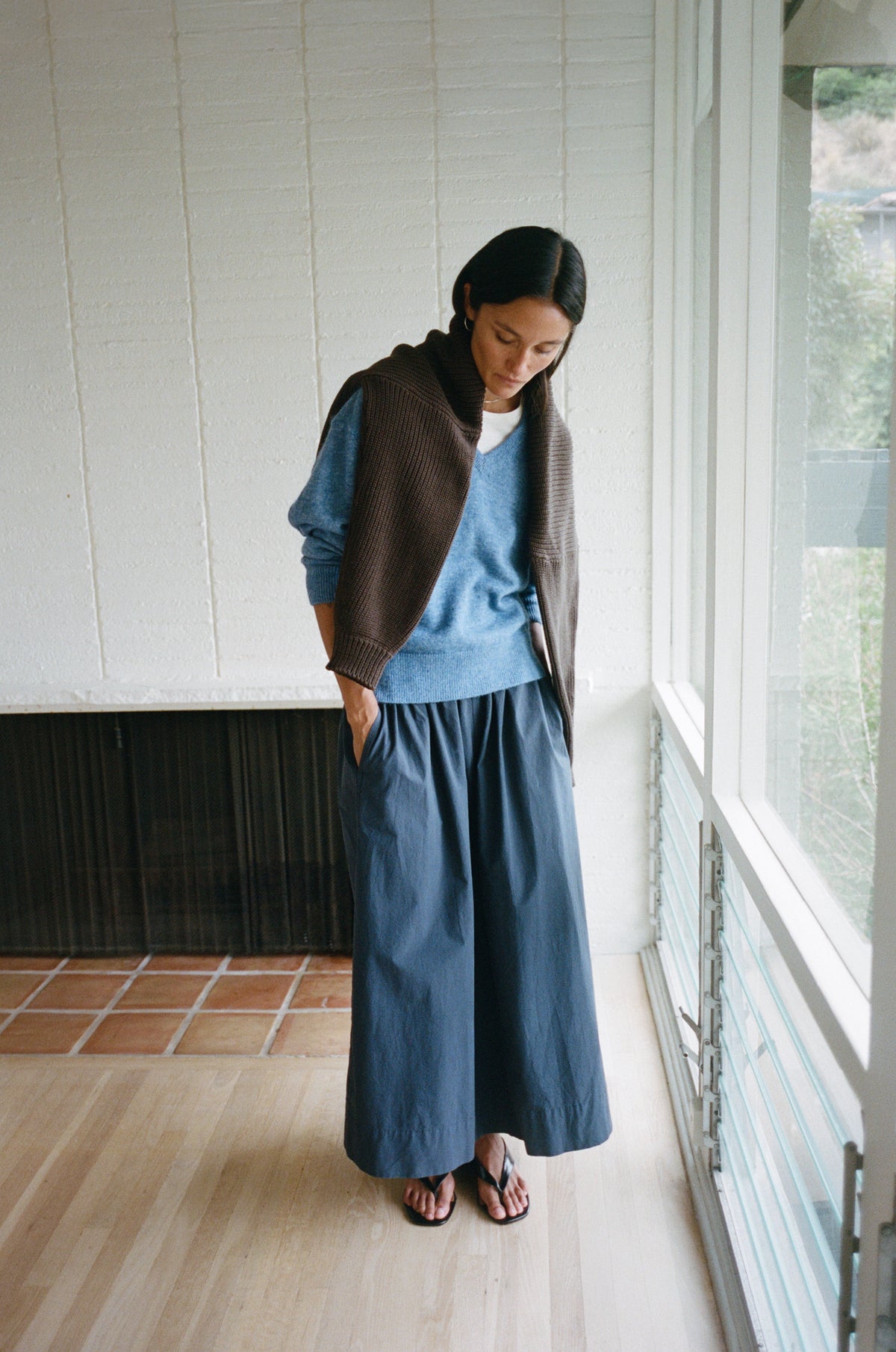 A woman stands indoors near a window wearing a blue knit sweater, a long blue skirt, and a brown cardigan draped over her shoulders, with sandals on a light wood floor.