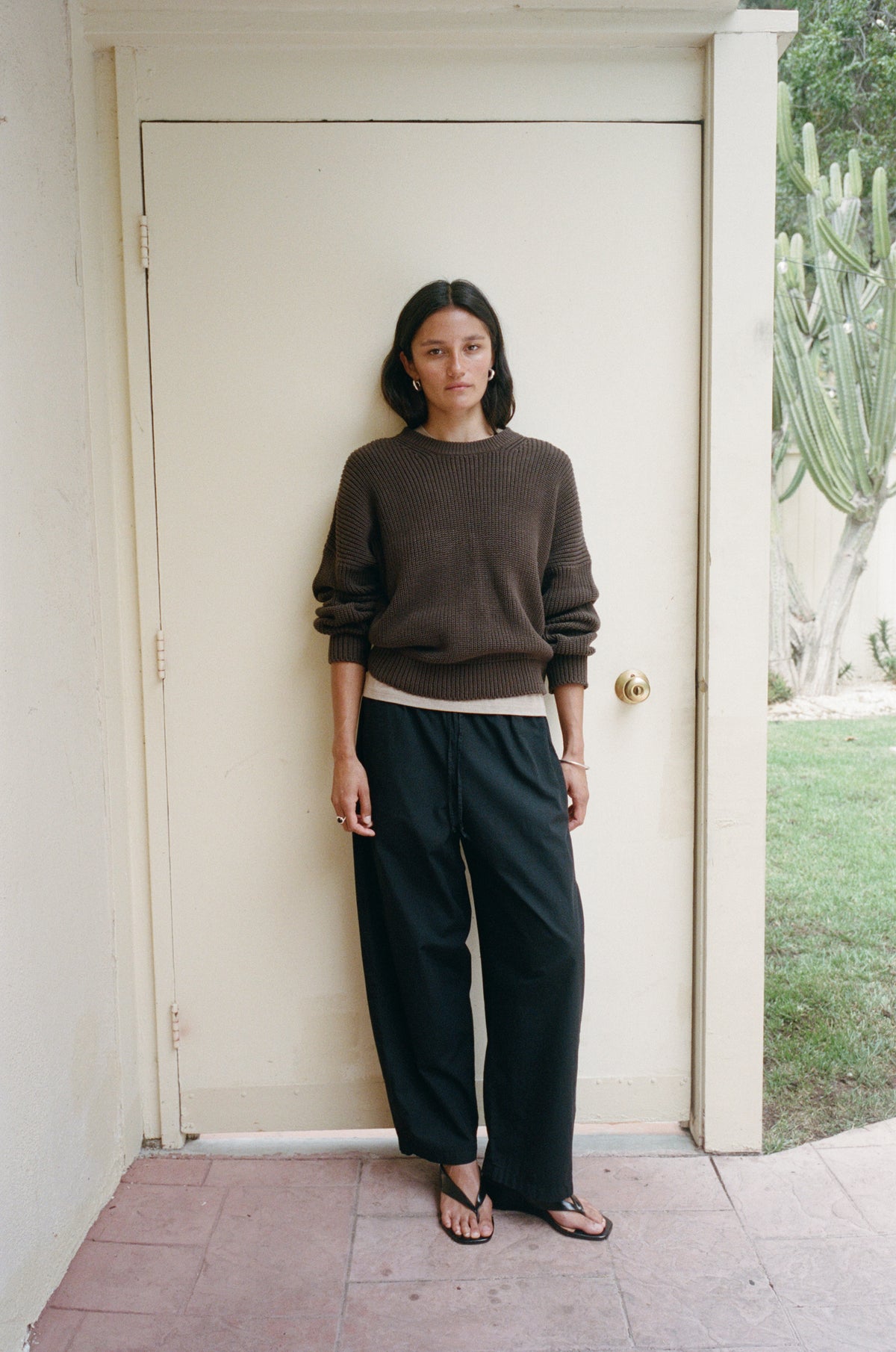 A woman stands in front of a cream-colored door wearing a dark brown crewneck sweater, black wide-leg pants, and black sandals.