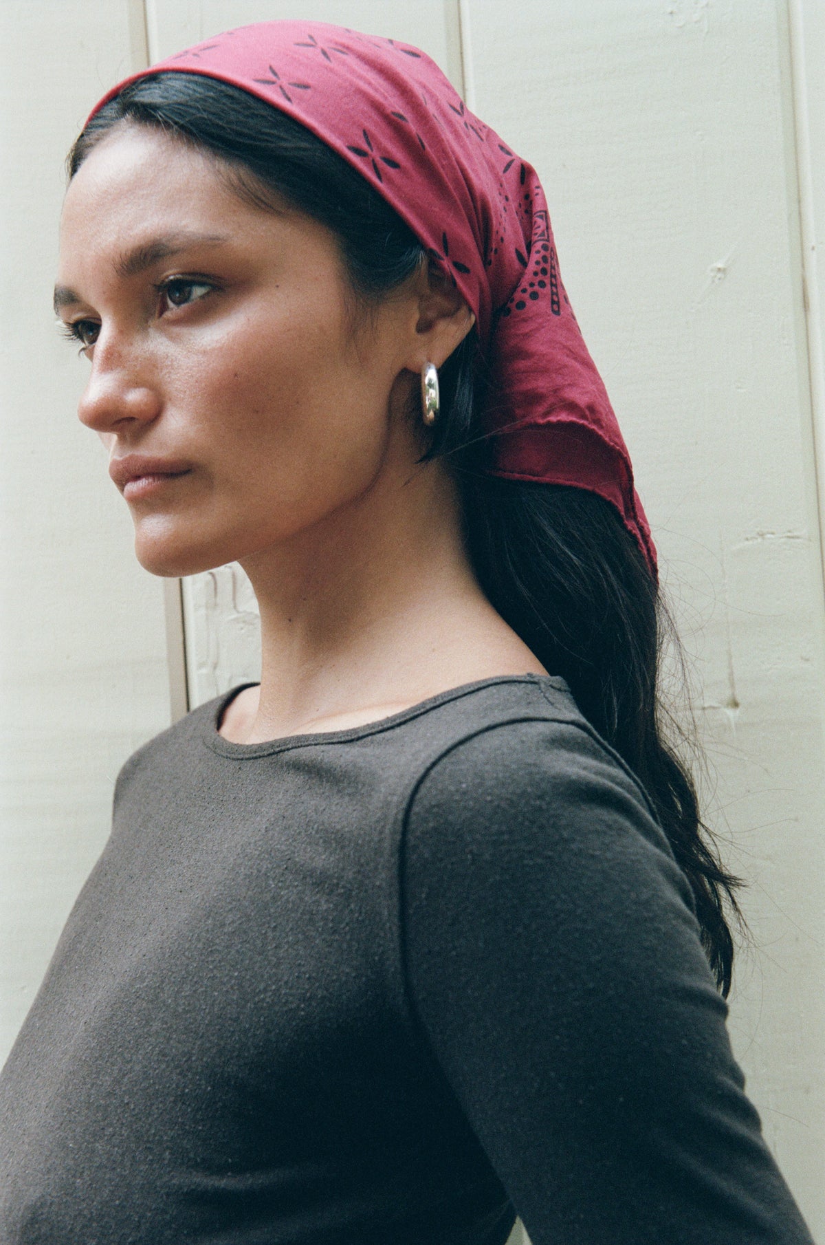 A woman in profile wearing a dark gray long-sleeve top and a dusty rose red patterned headscarf, standing against a light-colored wooden wall.