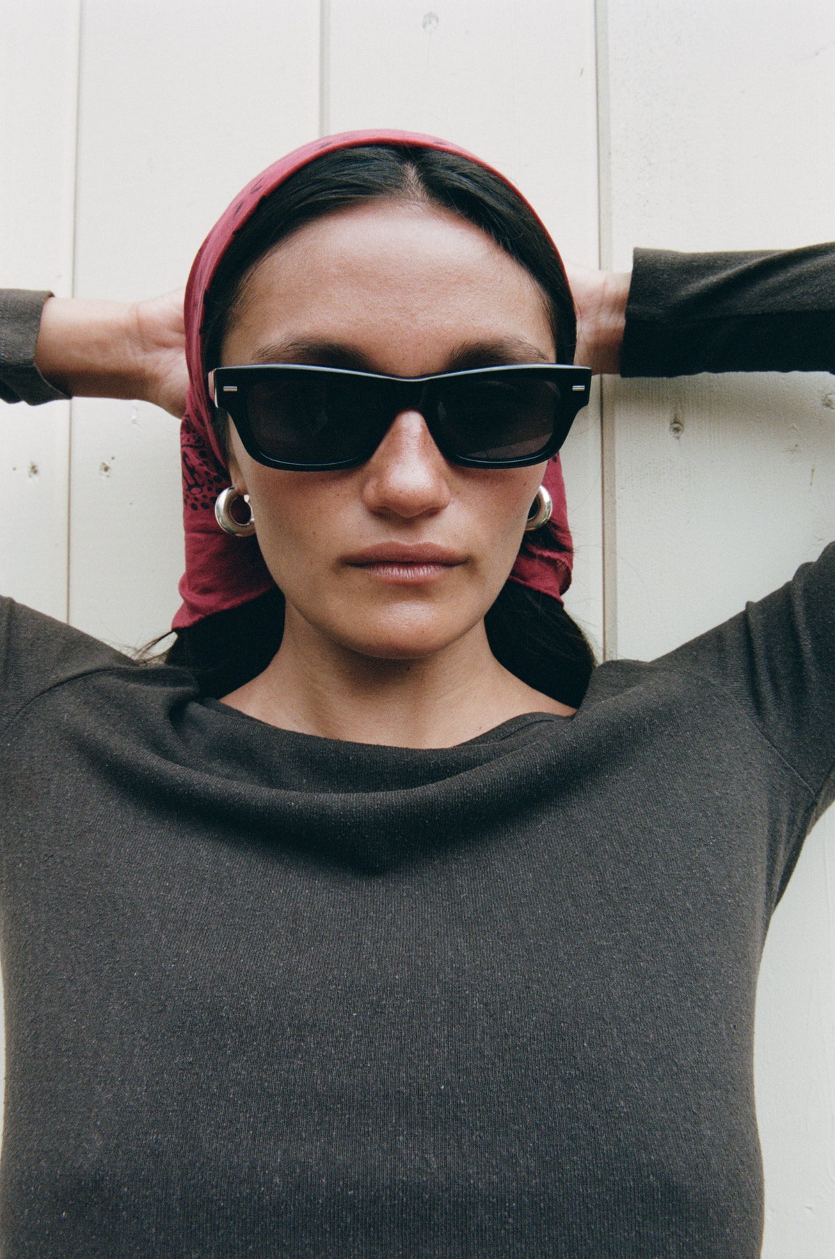 A woman wearing black cat-eye sunglasses, a dark charcoal cowl-neck sweater, and a red patterned headscarf, posing with her hands behind her head against a white paneled wall.