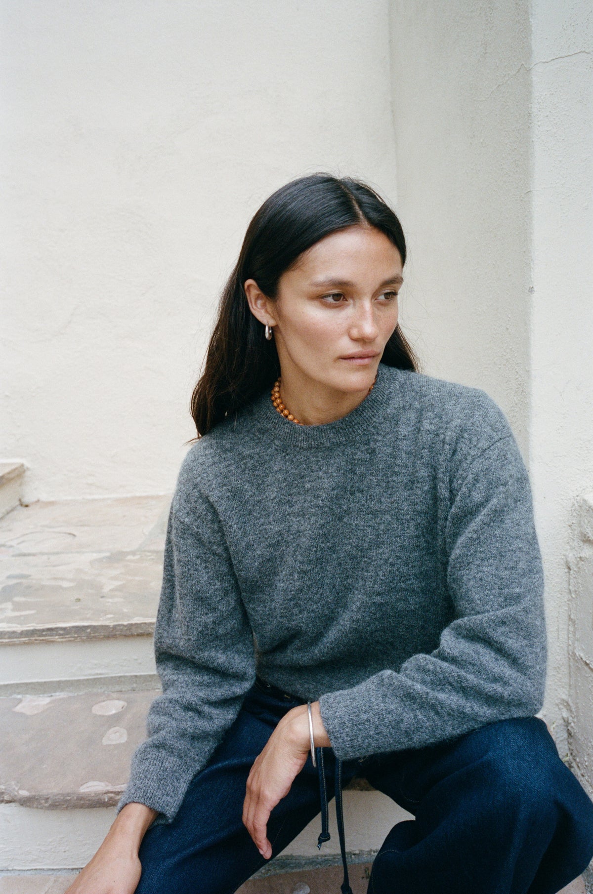 A woman with long dark hair sits on stone steps wearing a heathered gray knit sweater and dark blue pants against a light-colored stucco wall.