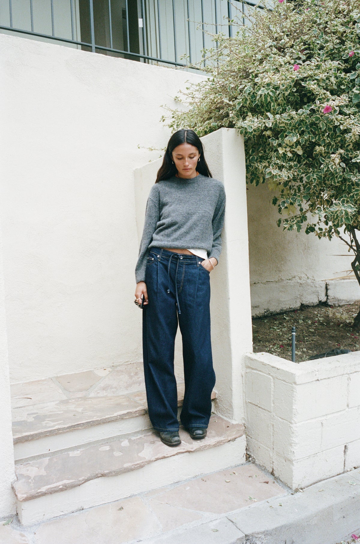 A woman stands on outdoor stone steps wearing a gray knit mock-neck sweater, dark wide-leg blue jeans, and black shoes, with a white wall and greenery in the background.