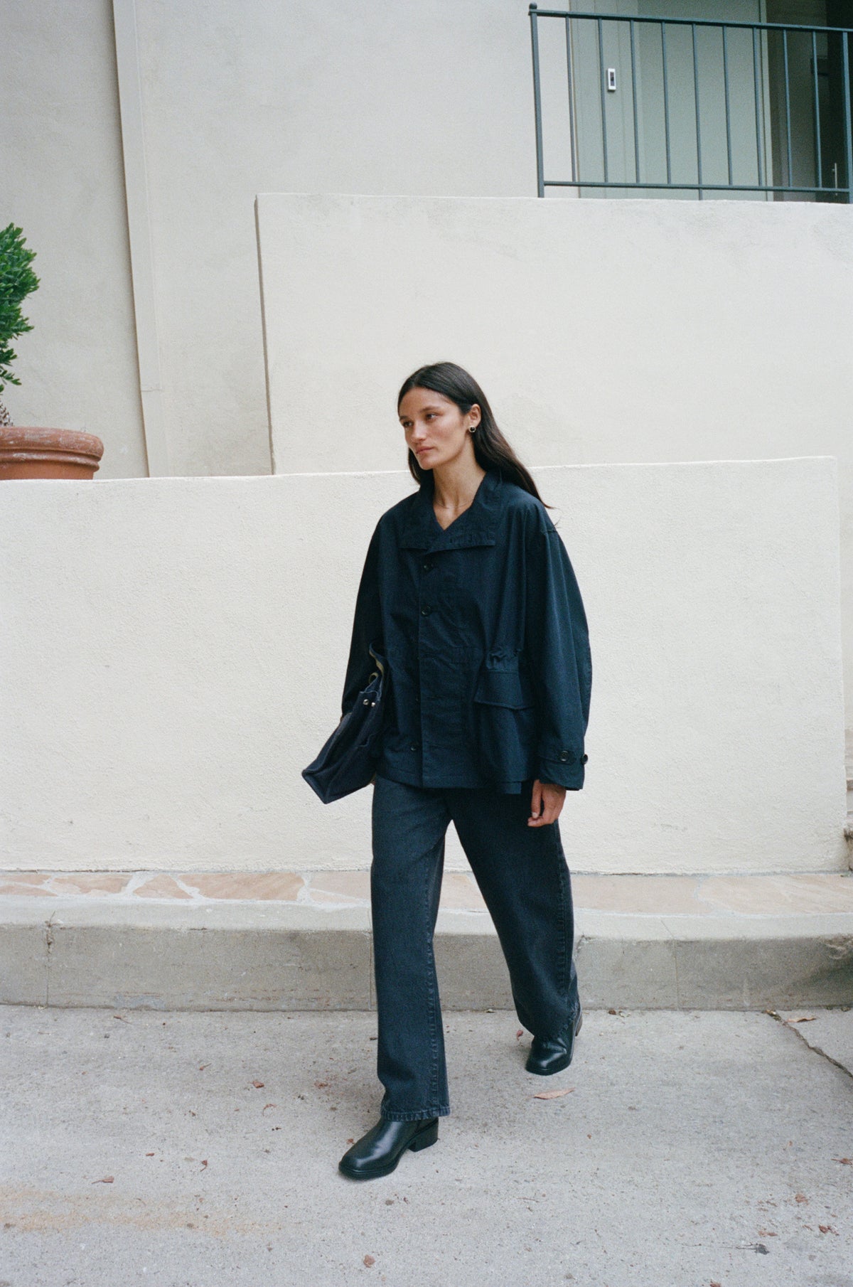 A woman wearing a dark navy blue oversized jacket and matching wide-leg trousers walks outdoors against a light beige stucco wall.