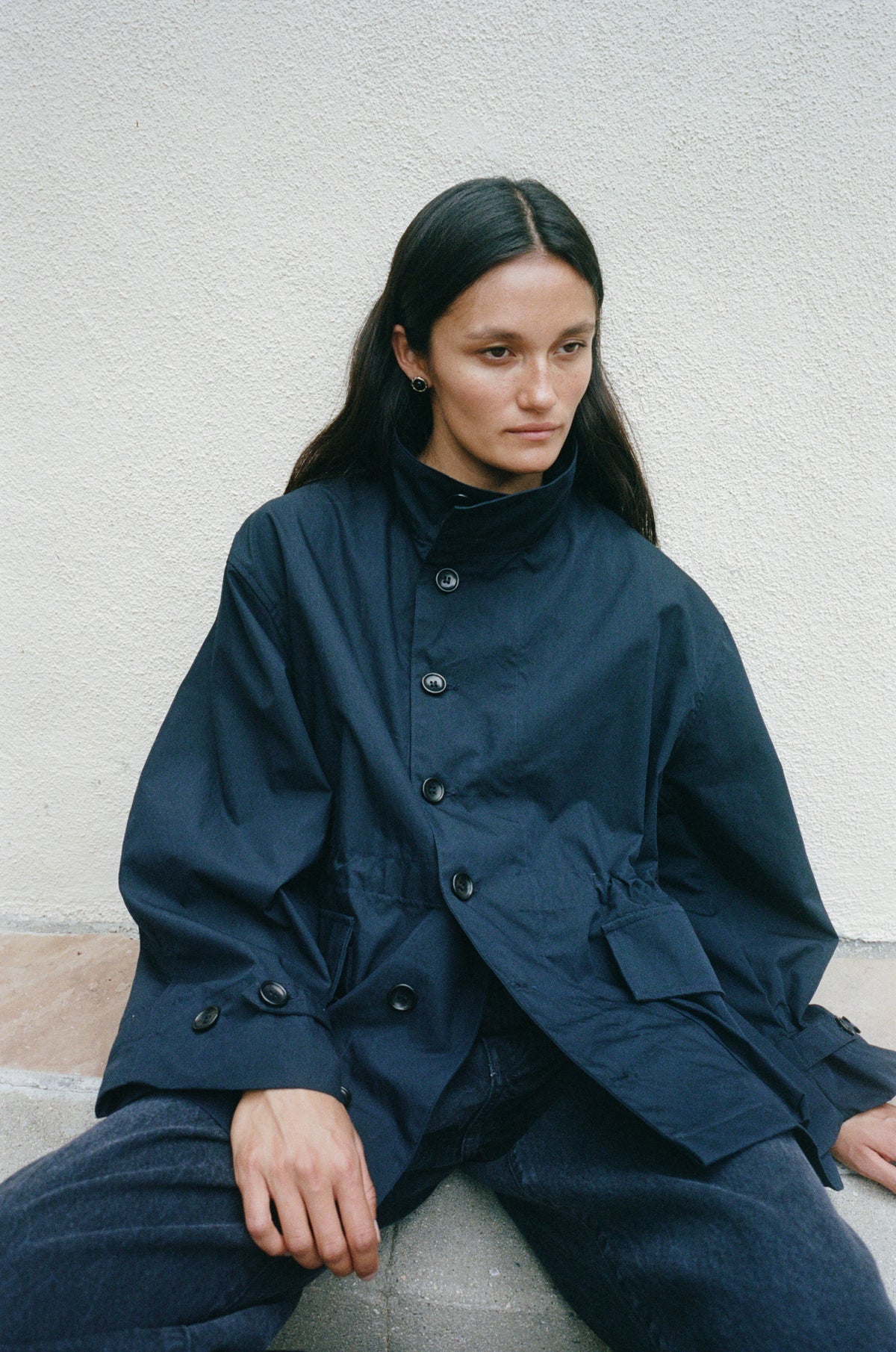 A woman with long dark hair sits against a textured off-white wall wearing a dark navy blue high-neck button-front jacket with oversized sleeves and matching dark pants.