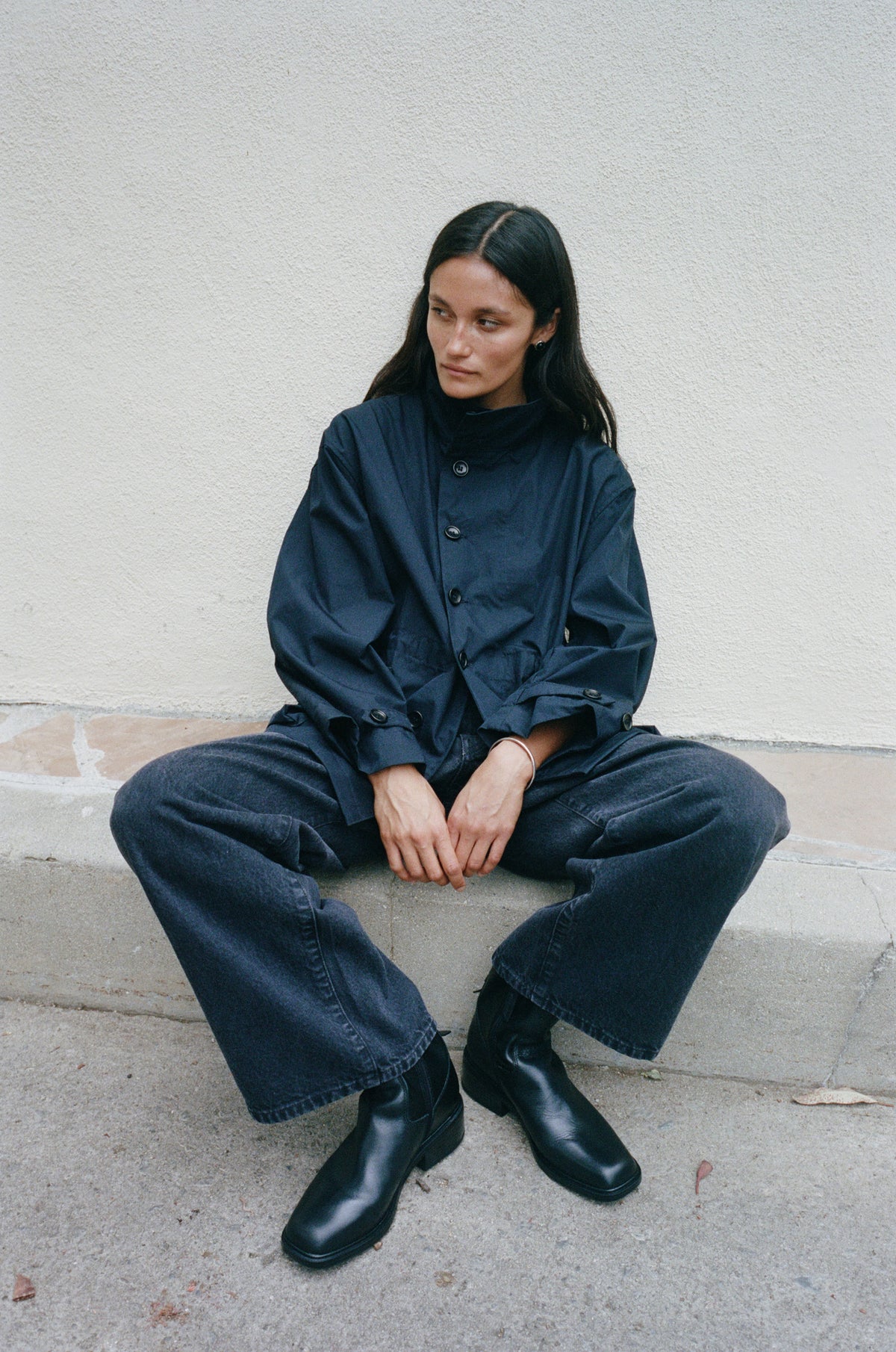 A woman sits on a concrete ledge against a textured off-white wall, wearing a dark navy blue high-neck button-up jacket, matching wide-leg pants, and black leather boots.