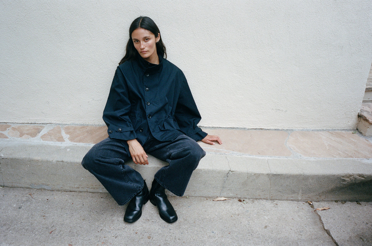 A model sits on a concrete ledge against a textured off-white wall, wearing a dark navy blue oversized button-up shirt and matching wide-leg pants with black boots.