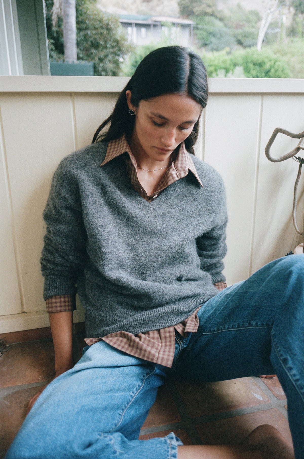A woman sitting on a tiled porch wearing a gray knit sweater over a brown plaid collared shirt and blue jeans.
