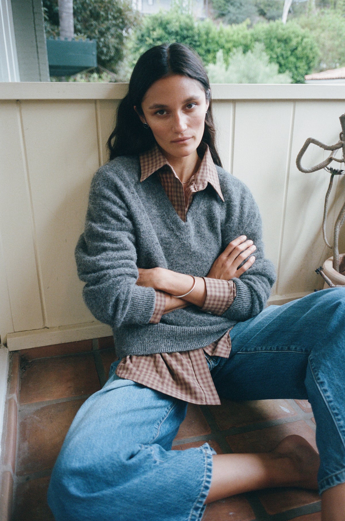 A woman sits on a porch wearing a gray knit sweater over a brown plaid button-down shirt, paired with blue jeans.