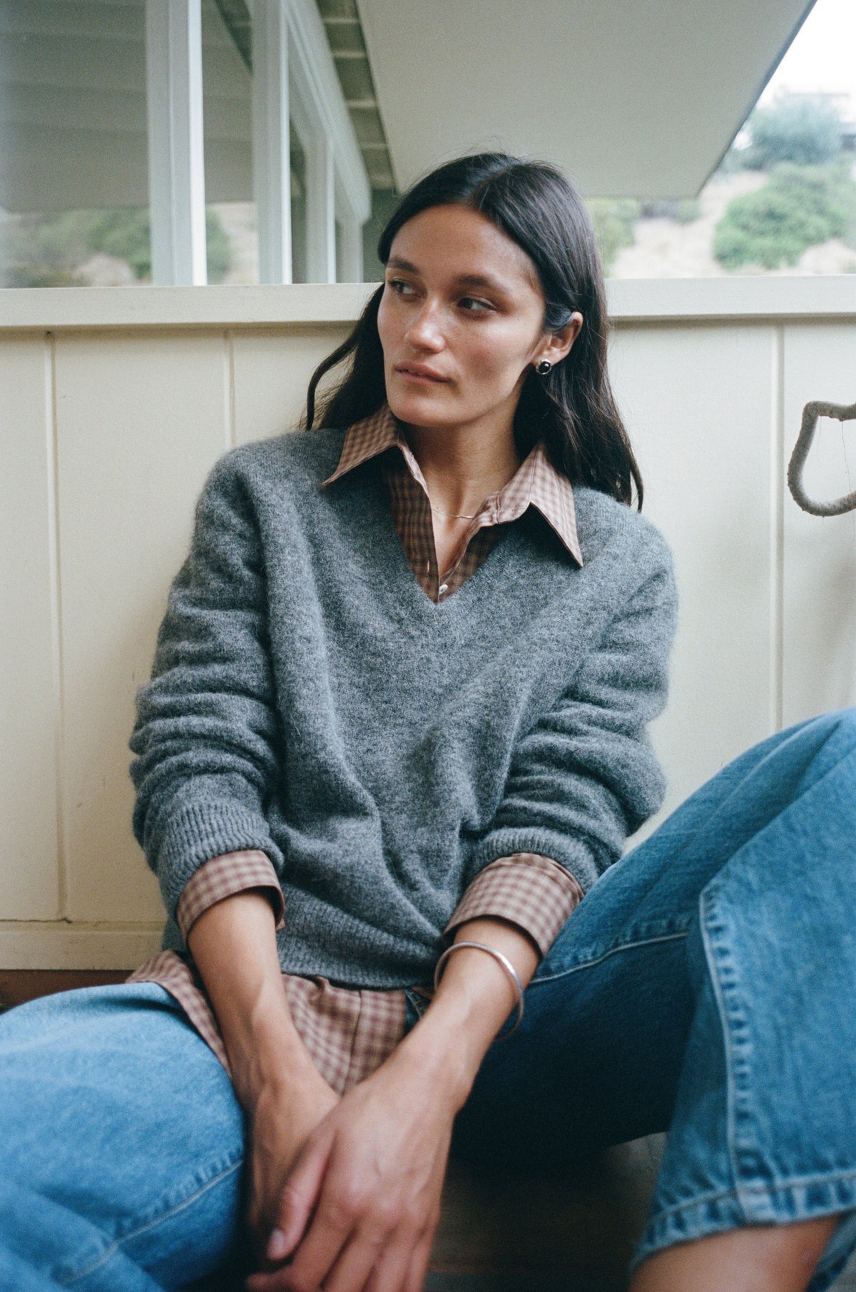 A woman sits on a porch wearing a gray knit sweater over a brown and beige plaid collared shirt, paired with blue jeans.