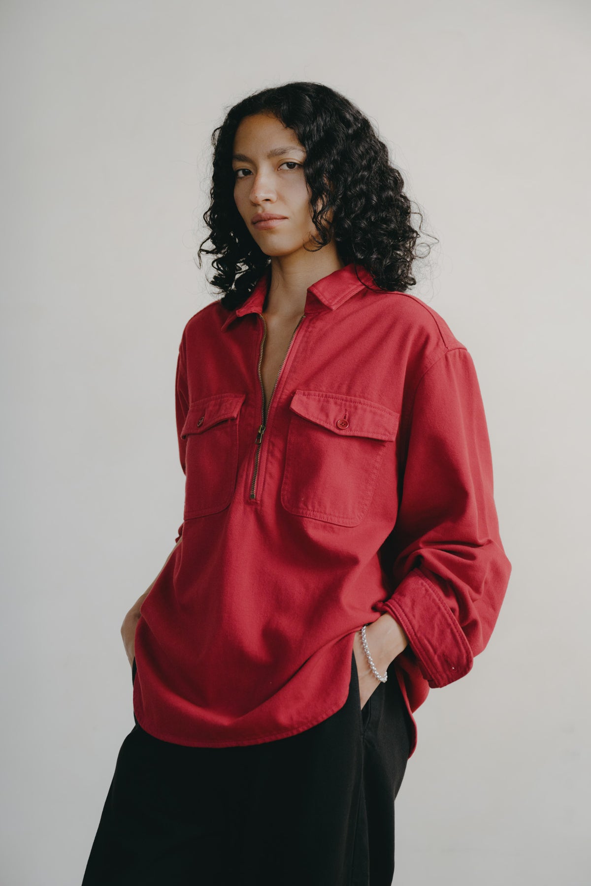 A model with curly dark hair wearing a red long-sleeve button-up shirt with a half-zip chest and black pants, standing against a plain light gray background.