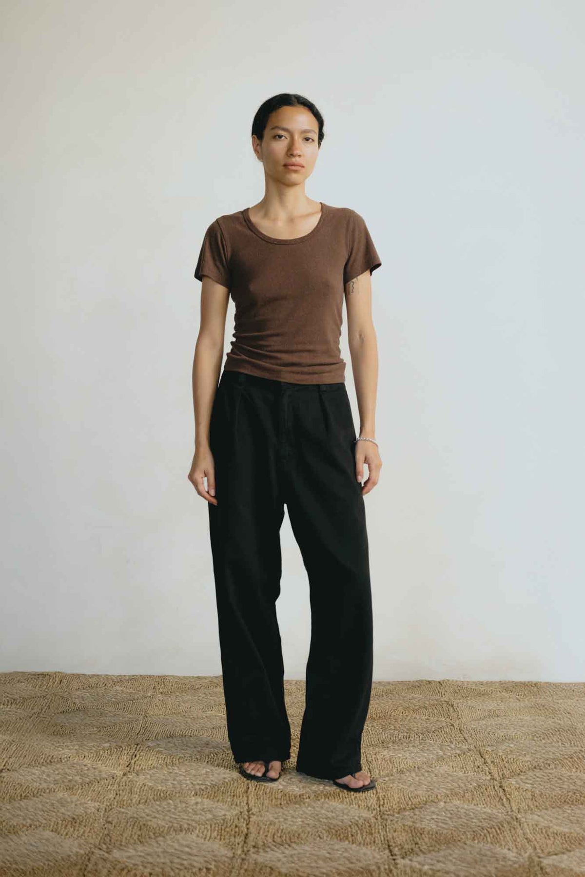 A model stands in a minimalist studio setting wearing a brown short-sleeve crewneck t-shirt and black wide-leg trousers, paired with black sandals on a textured beige rug.