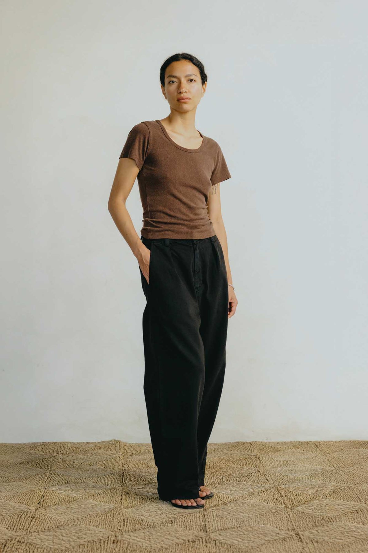 A woman stands in a studio wearing a fitted brown short-sleeve top and loose black wide-leg pants, with her hands in the pants pockets. The background is a light neutral wall with a woven beige floor.