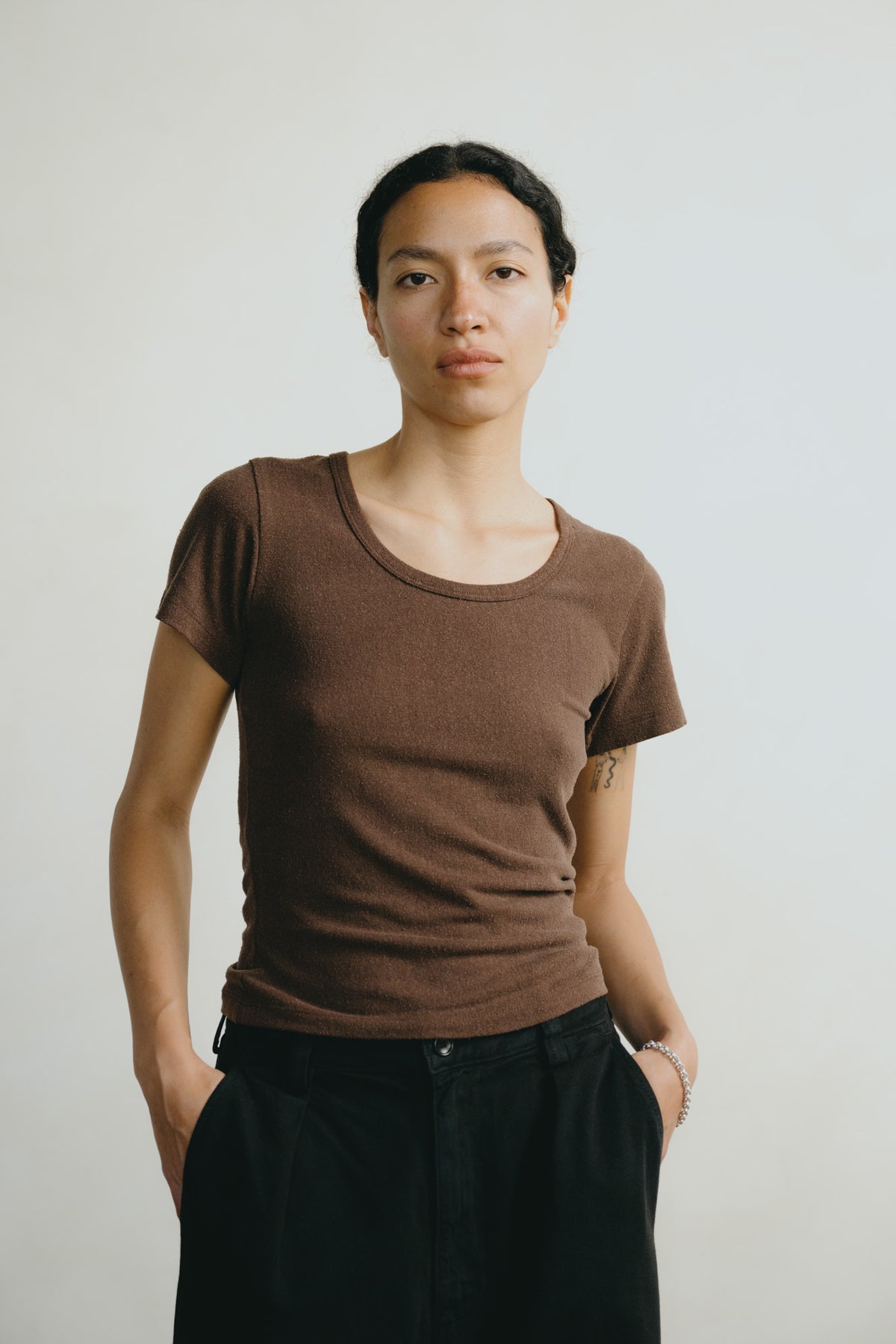 A woman wearing a fitted brown short-sleeve scoop-neck t-shirt and black pants, standing against a plain light gray background.