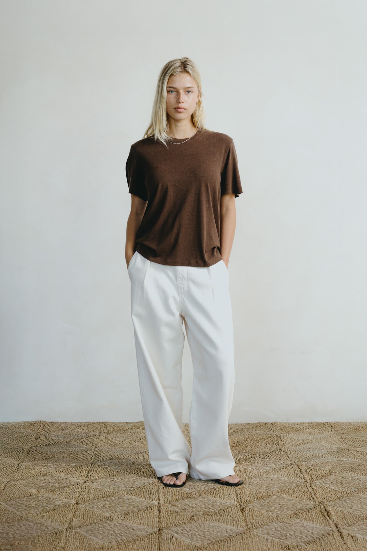 A woman stands against a plain off-white wall wearing a loose brown short-sleeve t-shirt and wide-leg white pants, with black sandals on a woven jute floor.