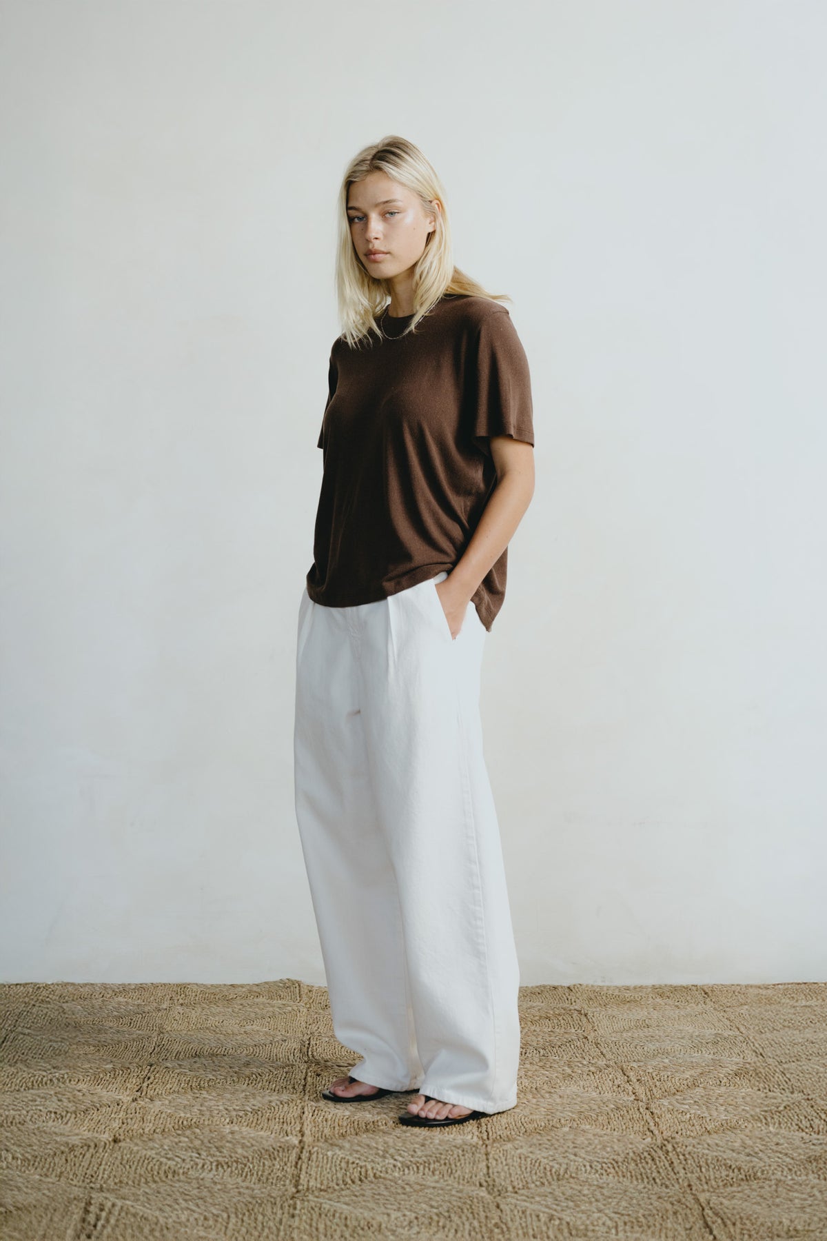 A woman stands in a minimalist room wearing a brown short-sleeve t-shirt and wide-leg white trousers, paired with black sandals.