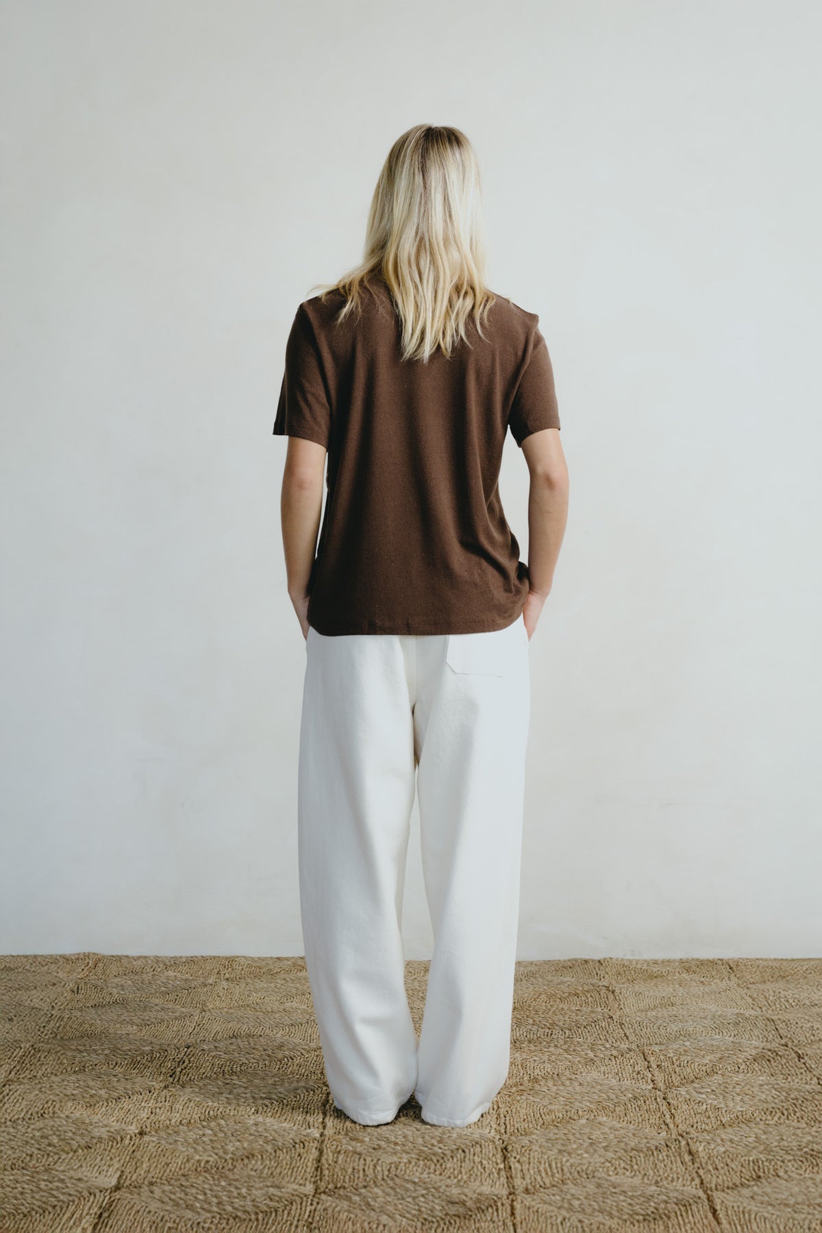 A woman with blonde hair is shown from behind, wearing a brown short-sleeve t-shirt and white wide-leg pants, standing on a woven straw rug against a plain off-white wall.