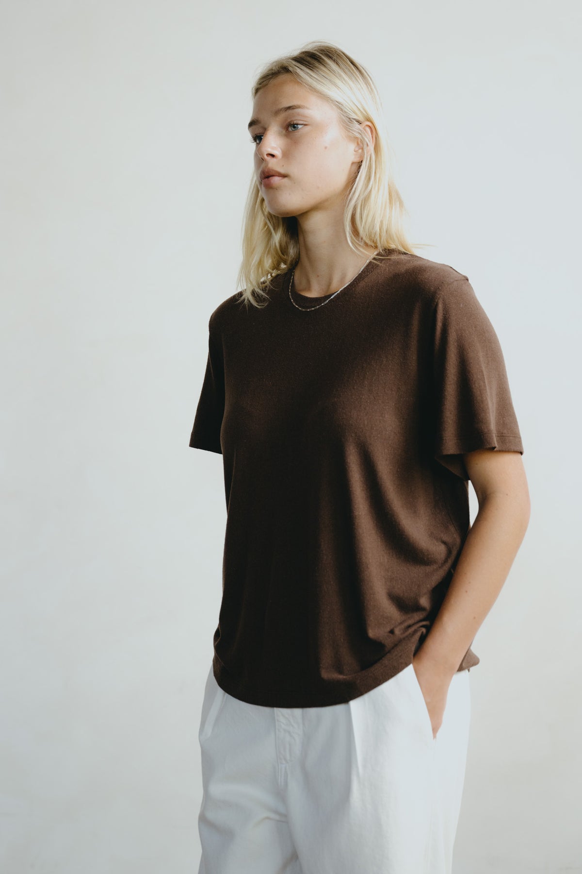 A woman wearing a dark brown short-sleeve crewneck t-shirt and white trousers, standing against a light gray studio background.
