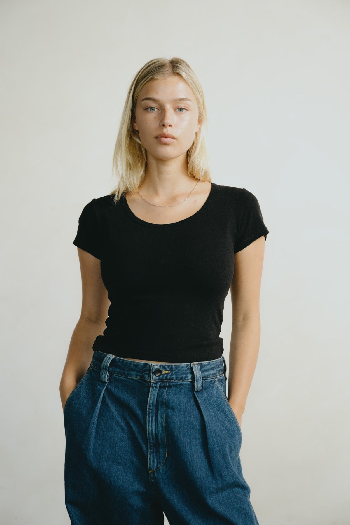 A blonde woman wearing a fitted black short-sleeve scoop-neck t-shirt and high-waisted blue jeans, standing against a plain light gray studio background.