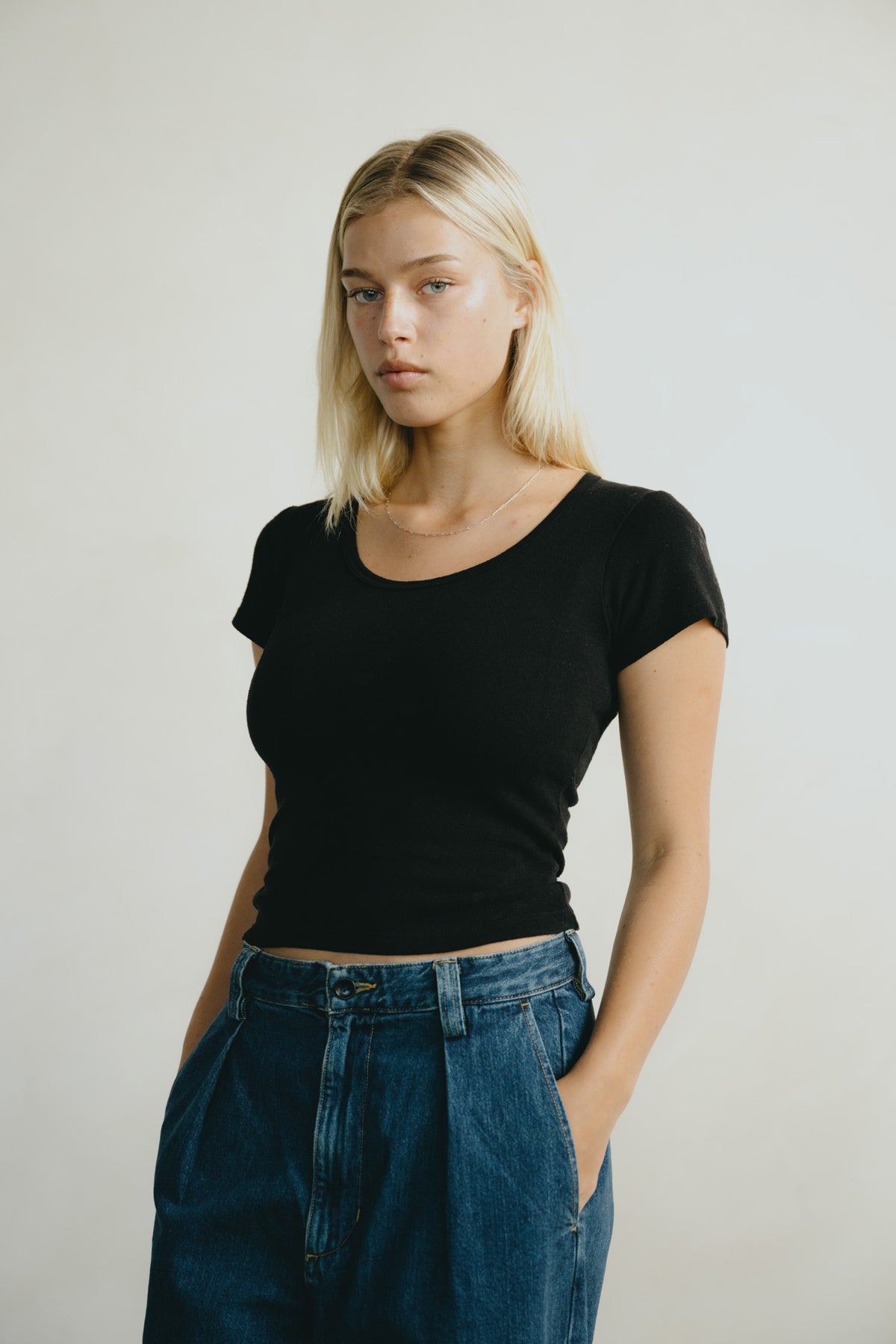 A blonde woman wearing a black short-sleeve scoop-neck t-shirt and high-waisted blue jeans, standing against a plain light gray studio background.